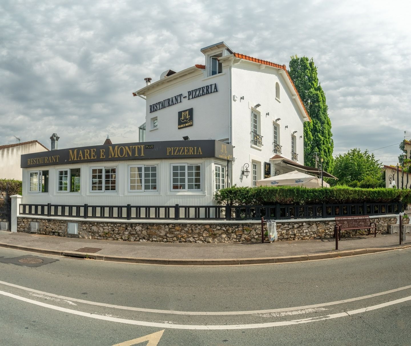 Bâtiment blanc d'un restaurant en bord de route, avec une enseigne noire et un mur de pierre, sous un ciel nuageux.