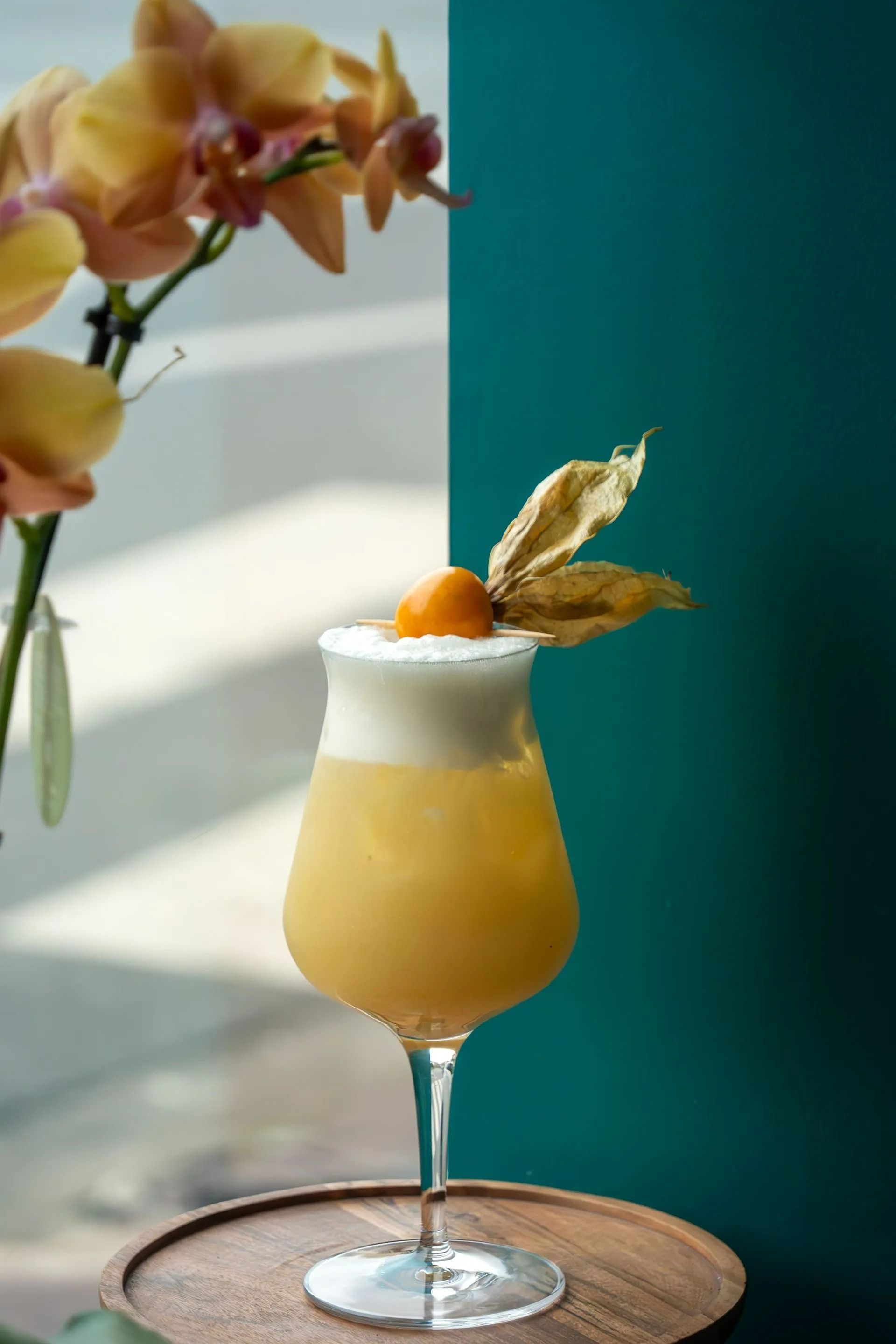 Un cocktail jaune servi dans un verre à pied, coiffé d'une mousse onctueuse et garni de physalis, sur une table en bois.