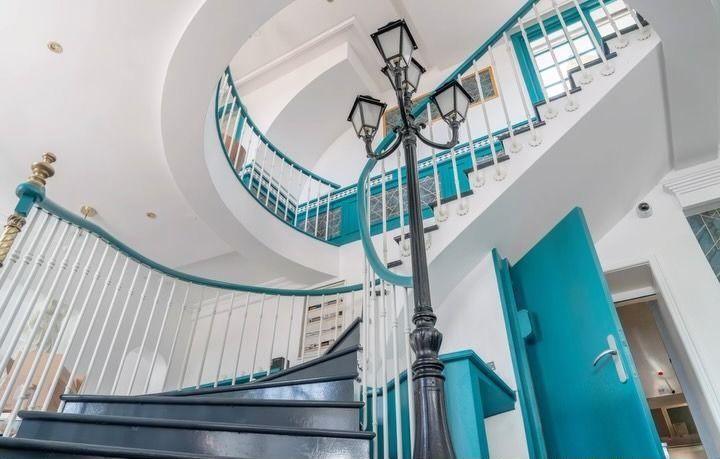 Escalier courbe, murs blancs, rampes turquoise et porte turquoise ouverte dans un intérieur lumineux.