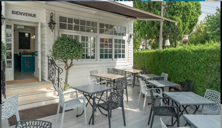 Terrasse de café en plein air avec tables et chaises en métal noir, à côté d'un bâtiment blanc et d'une haie verte.