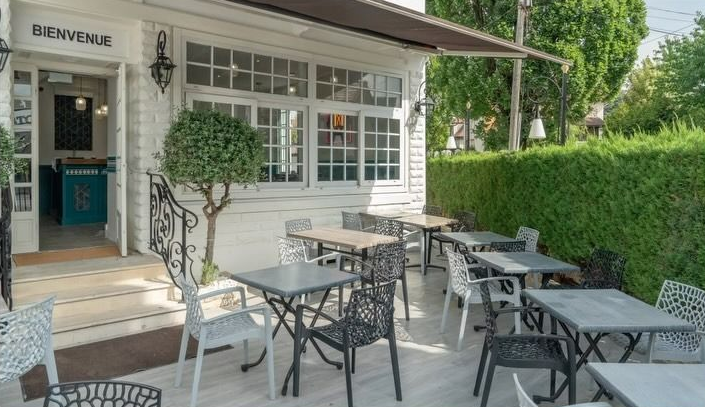 Terrasse de café en plein air avec tables et chaises en métal noir, à côté d'un bâtiment blanc et d'une haie verte.