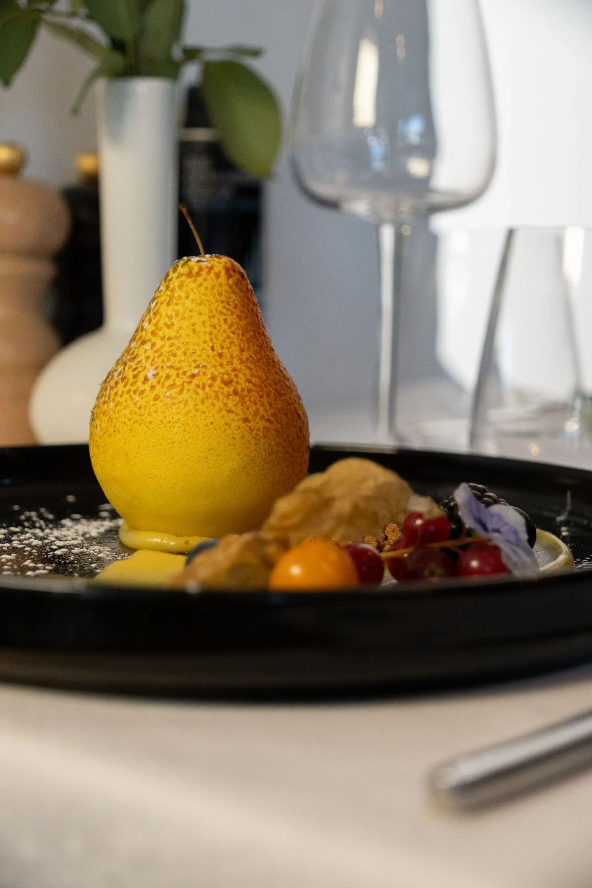 Dessert en forme de poire présenté sur une assiette noire, garni de fruits, avec un verre de vin en arrière-plan.