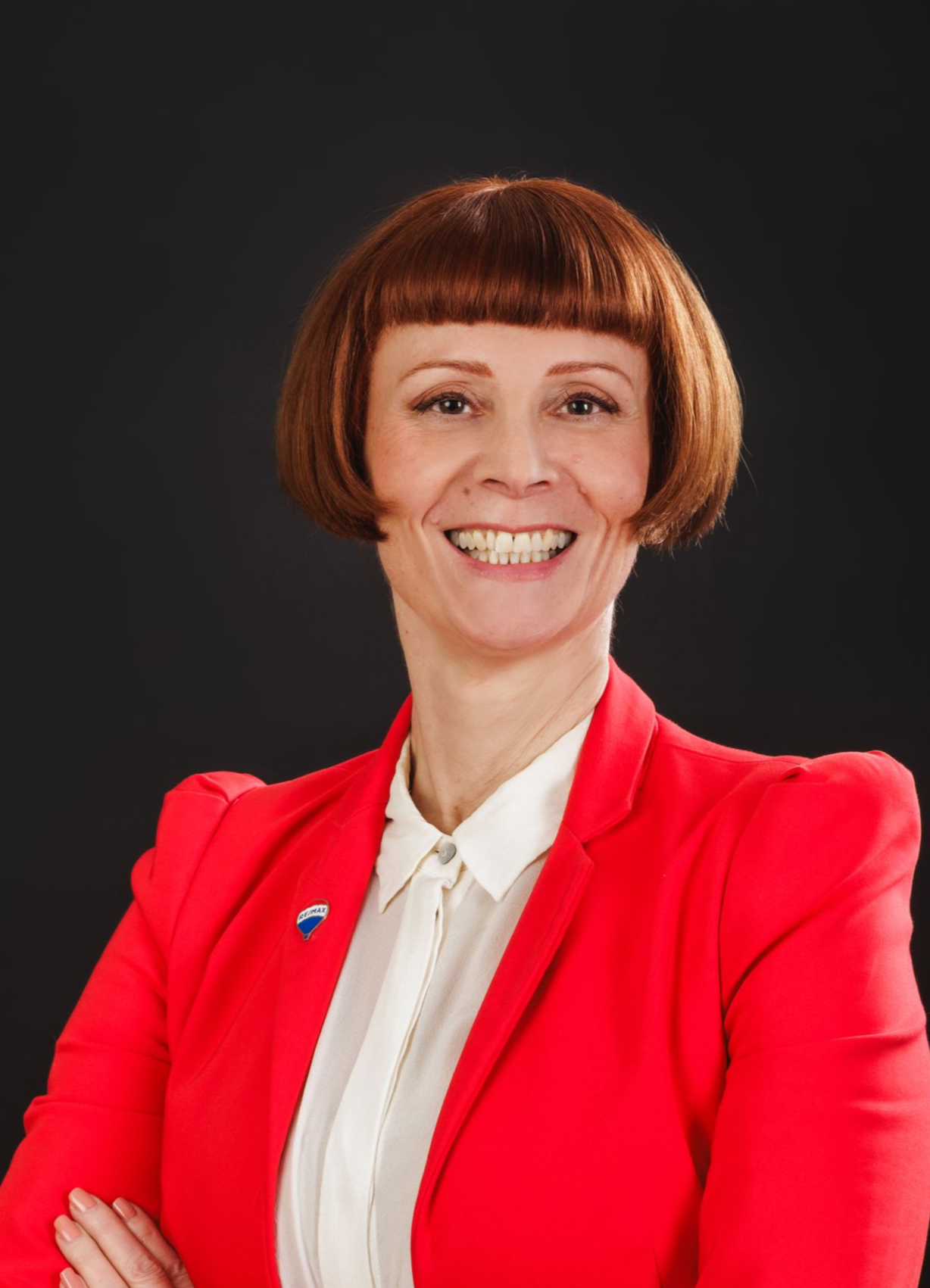 Frau mit rotem Bob-Haarschnitt, die einen roten Blazer und ein weißes Hemd trägt, lächelt mit verschränkten Armen.