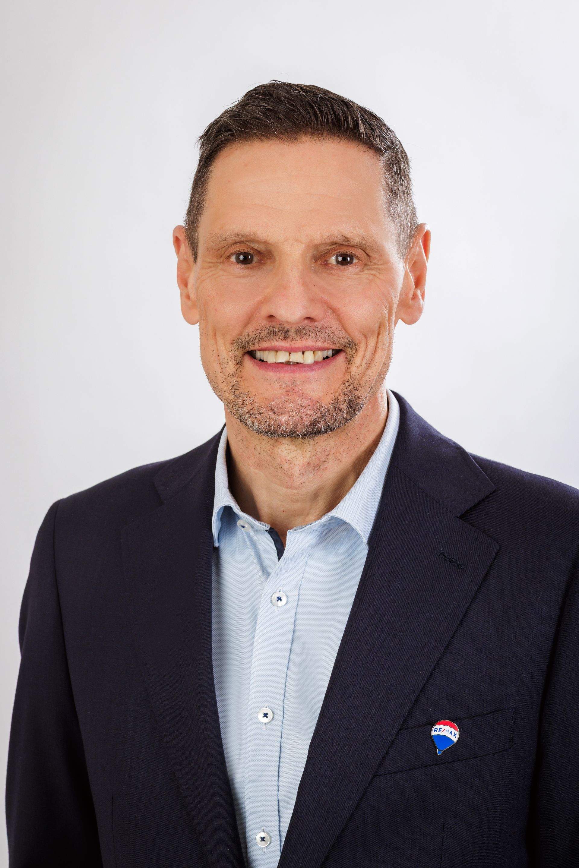 Mann im dunkelblauen Blazer und hellblauem Hemd, lächelnd, RE/MAX-Anstecknadel sichtbar.