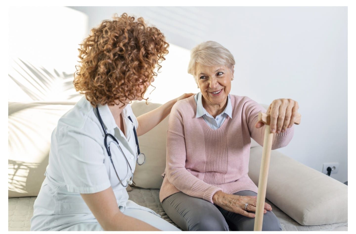 Cuidadora ayuda a una anciana a caminar al aire libre, sonriendo, tomándole la mano y el brazo.