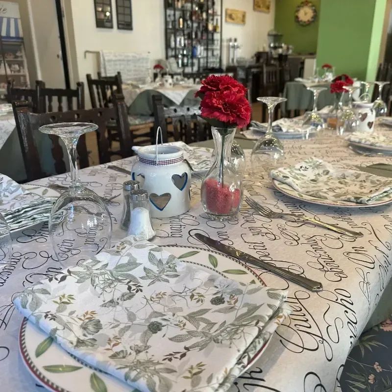 Una mesa de restaurante puesta para comer, con servilletas florales, una flor roja en un jarrón, cristalería