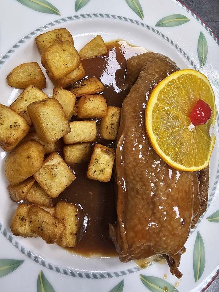 Pechuga de pollo asada servida en un plato con patatas en dados, salsa marrón y una rodaja de naranja como guarnición.