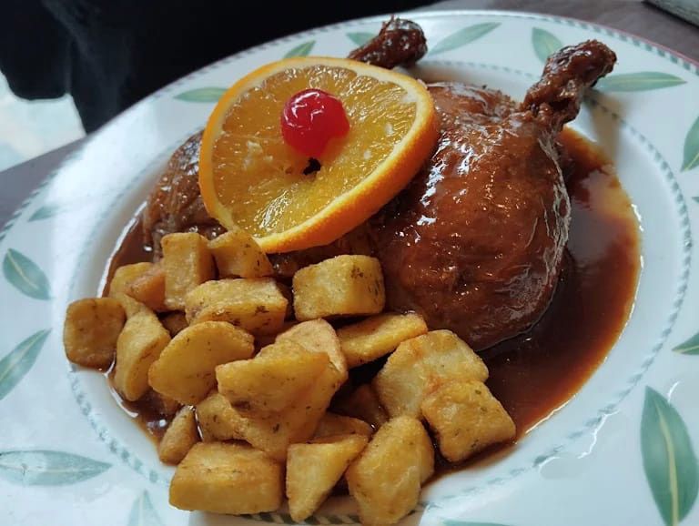 Pollo estofado con rodaja de naranja y cubos de patata asada en un plato blanco.