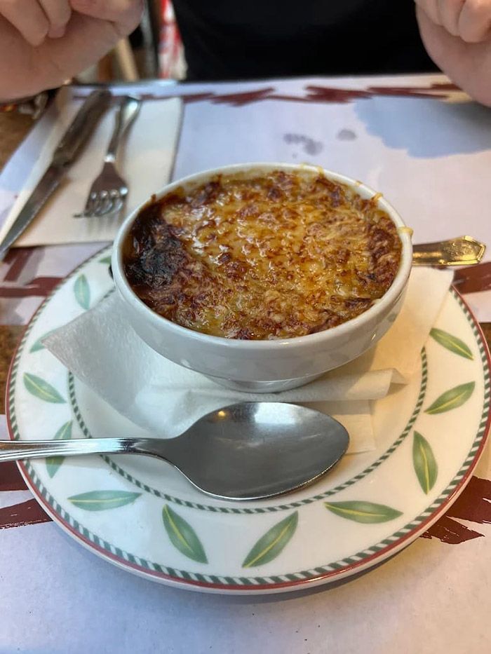 Crème brûlée en una taza blanca sobre un platillo estampado, con cuchara y tenedor sobre una mesa.