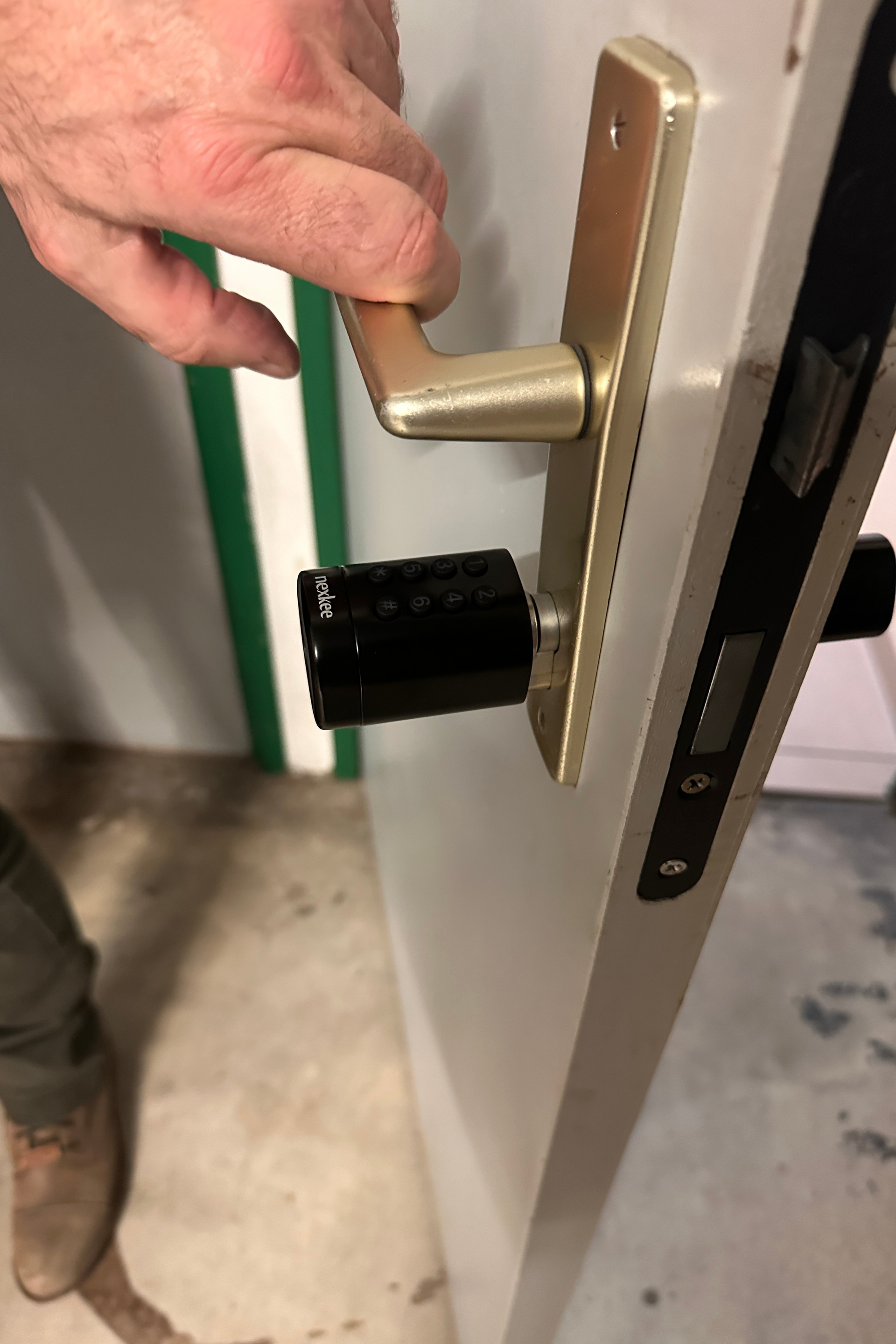 Main ouvrant une porte avec une poignée dorée et une serrure noire ; une personne portant des bottes marron est visible.