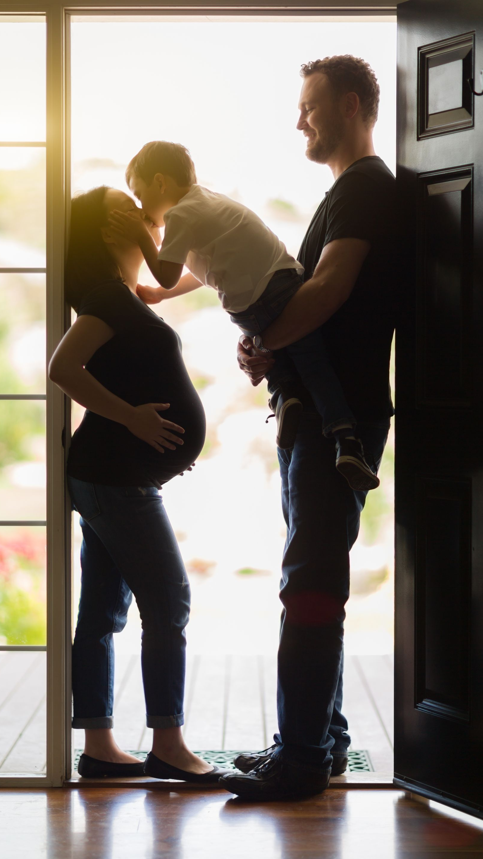 Une femme enceinte et un homme tiennent un enfant dans l'embrasure d'une porte, rétroéclairés par une lumière vive.