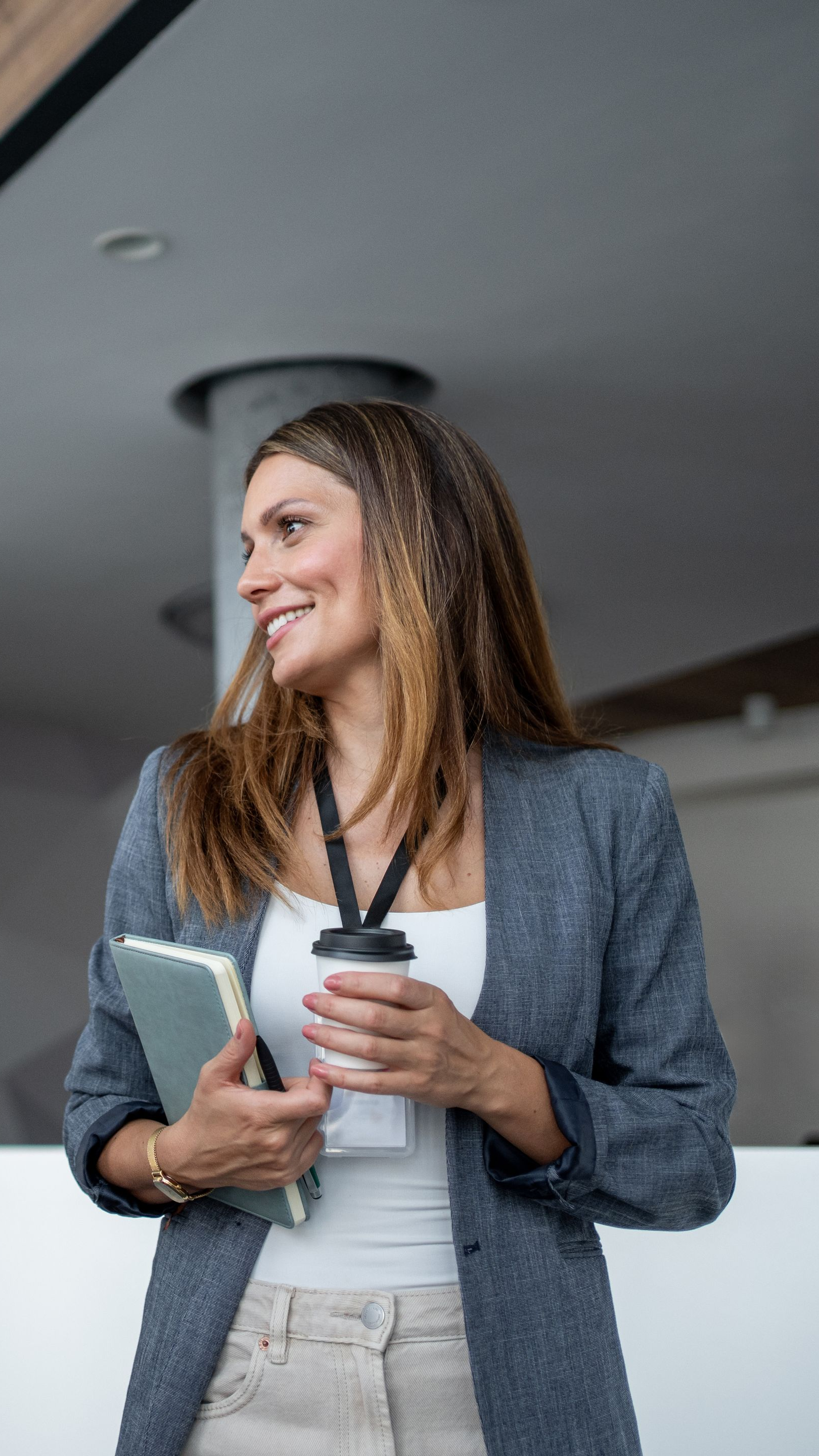 Une femme en blazer tient un café et un cahier, souriant, à l'intérieur d'un bâtiment moderne.