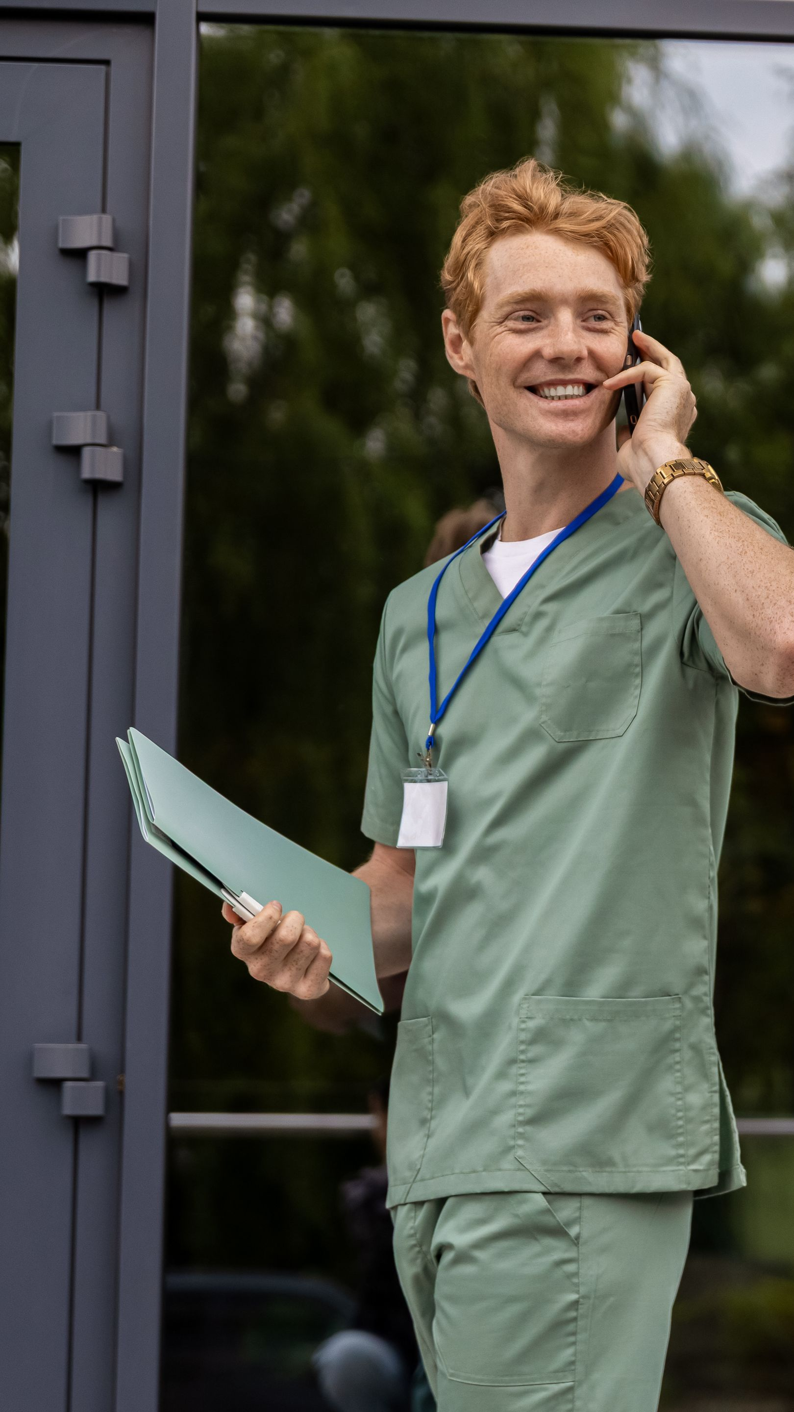 Un homme en tenue verte parle au téléphone, tient un presse-papiers, debout à l'extérieur d'un bâtiment.