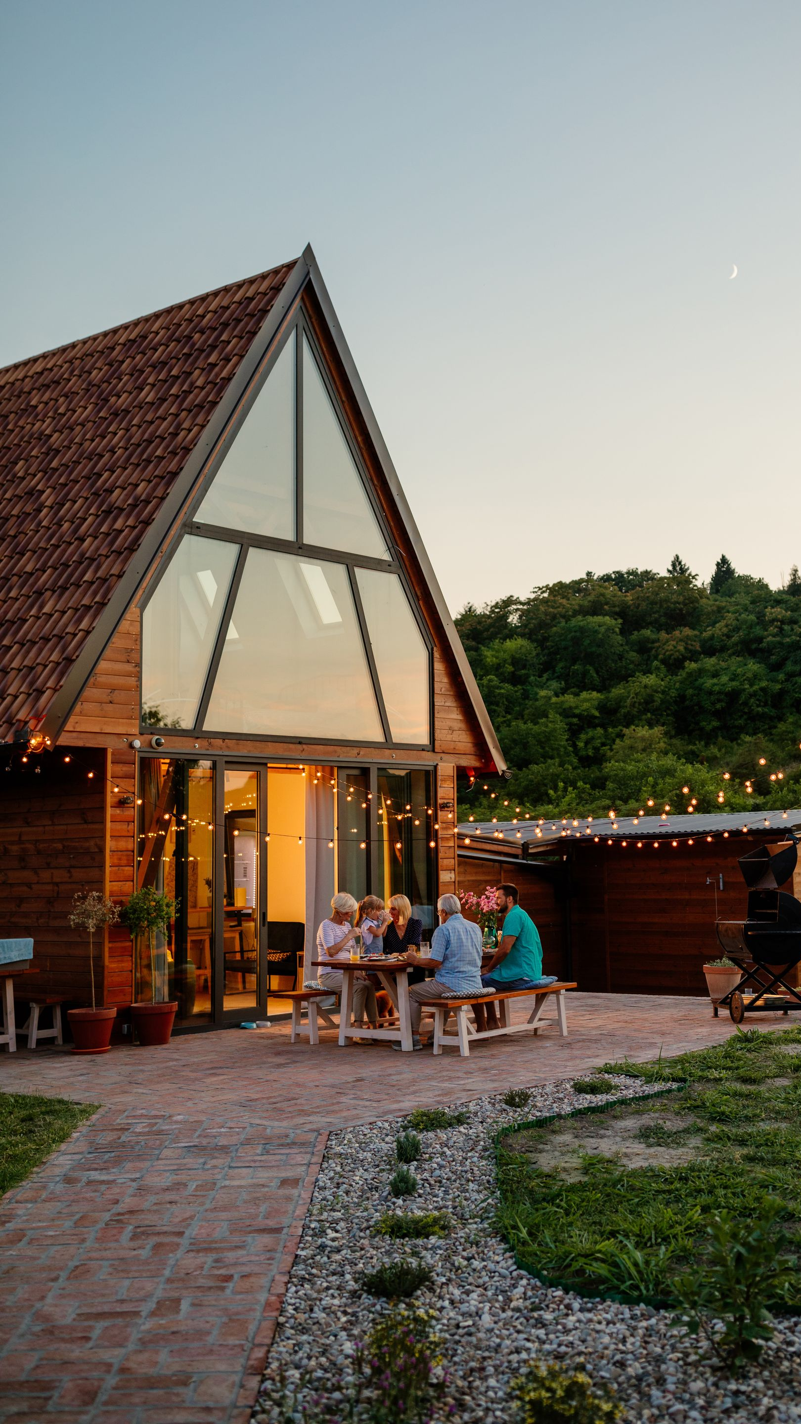 Des gens assis à une table à l'extérieur d'une maison en bois en forme de A, savourant un repas sous des guirlandes lumineuses.