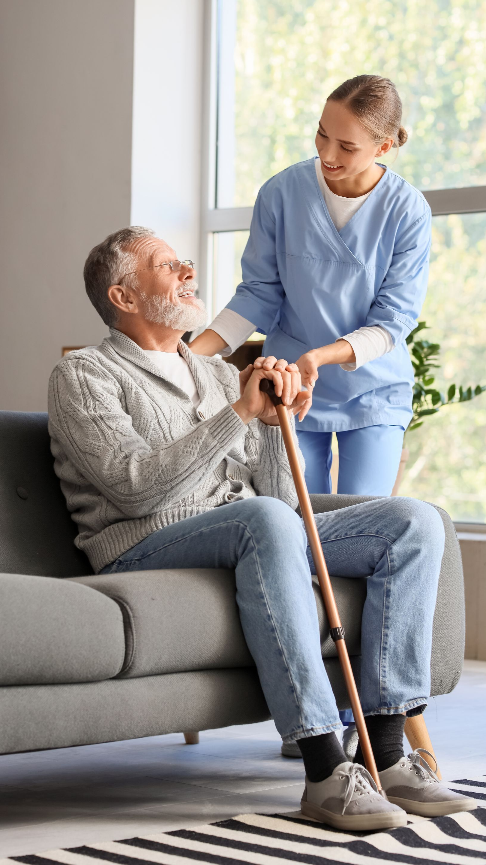 Homme âgé sur un canapé, soutenu par un gardien. Intérieur souriant et ensoleillé. Canne visible.