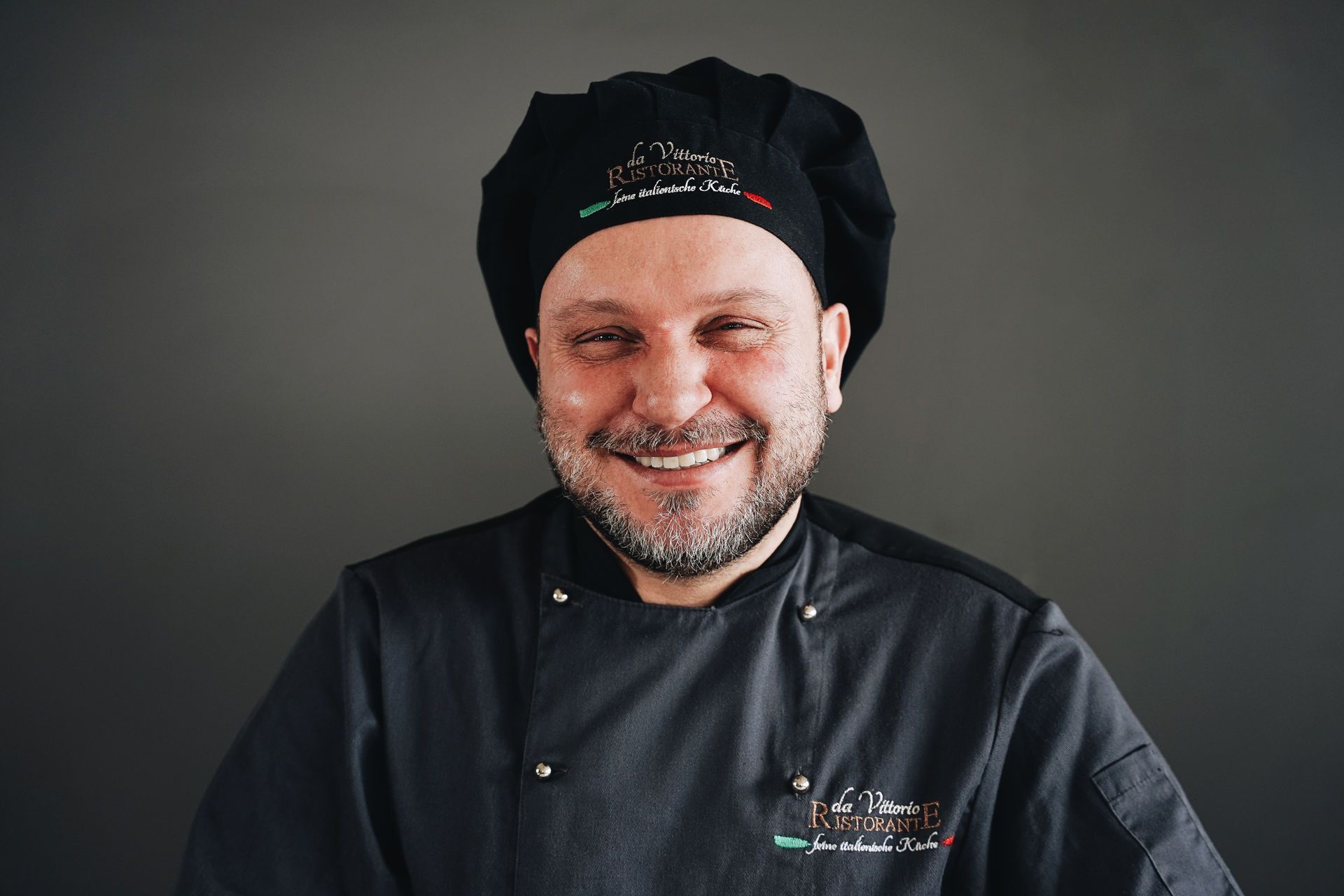 Koch mit Bart und Lächeln, trägt eine schwarze Kochmütze und Jacke vor einem grauen Hintergrund.