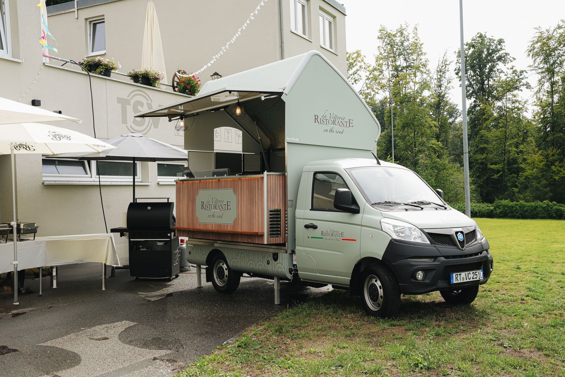 Ein hellgrüner Imbisswagen mit hölzerner Theke, der auf einer Kiesfläche neben einem Gebäude parkt, und ein Außentisch.