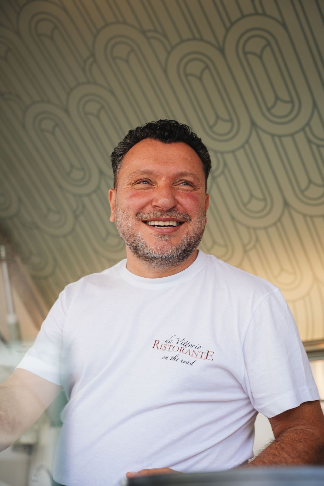 Eine Person mit Bart und weißem T-Shirt lächelt vor einem Hintergrund mit sich wiederholendem ovalen Muster.