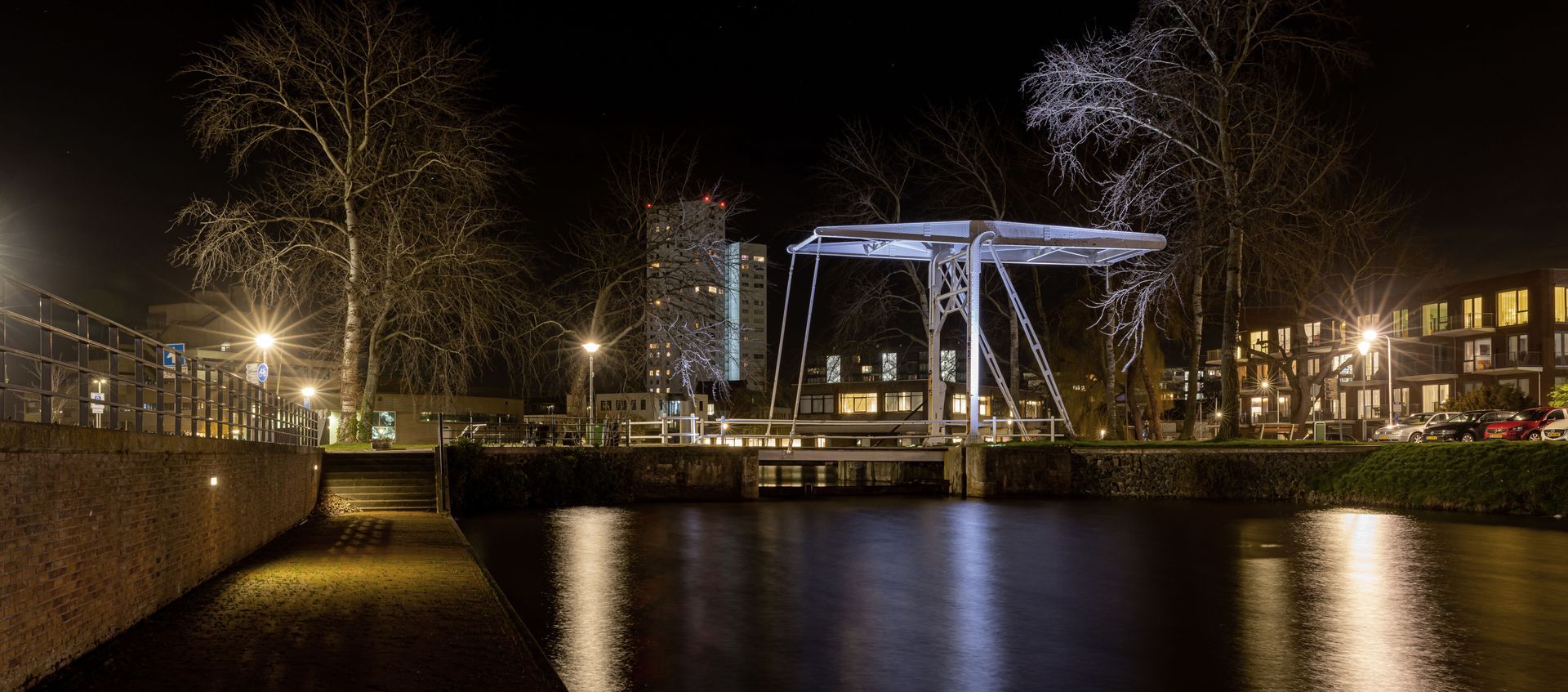 Nachtelijk uitzicht op een brug over een kanaal met verlichte gebouwen en bomen die in het water worden weerspiegeld.