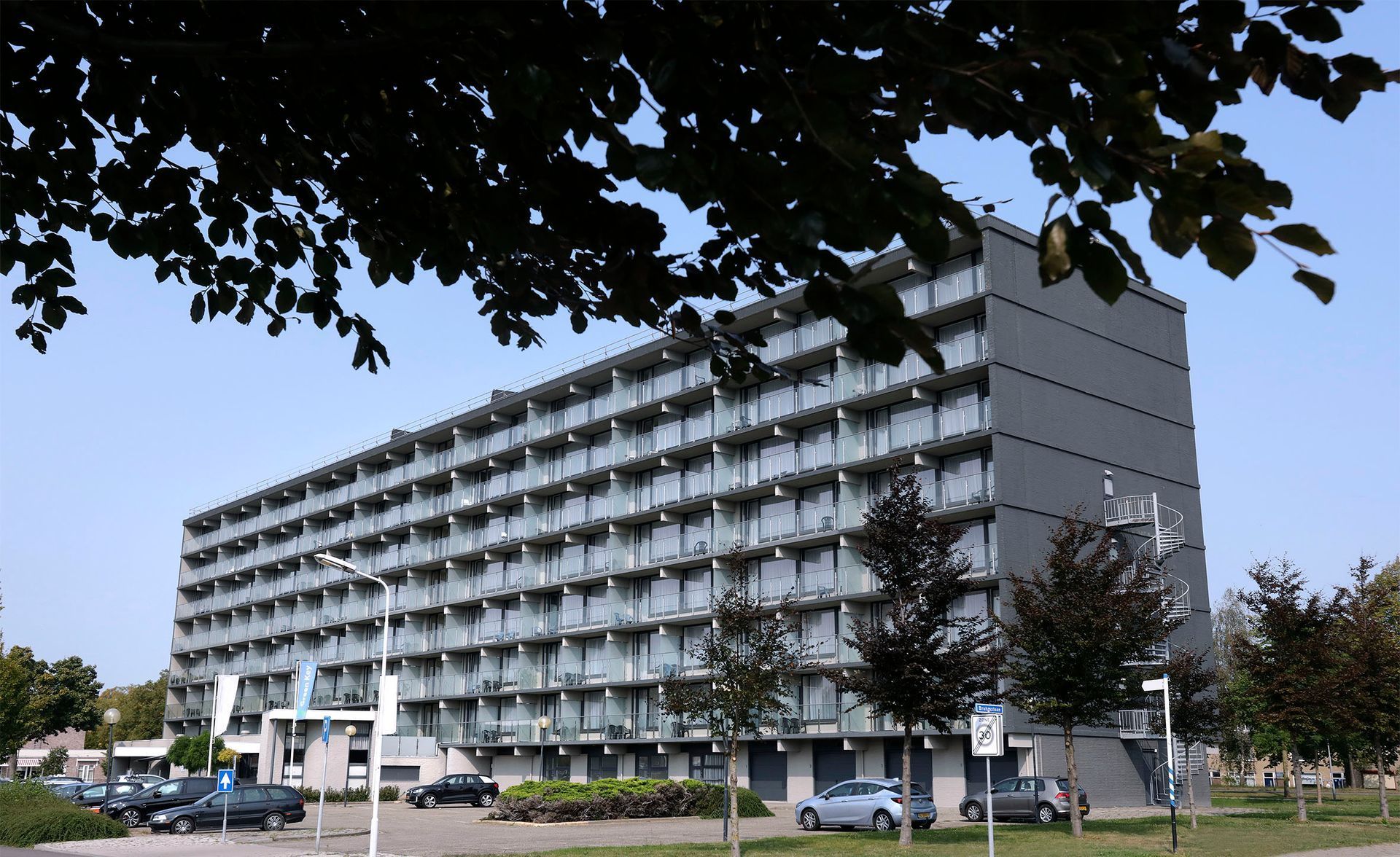Multi-story gray apartment building with balconies, trees in foreground, blue sky.