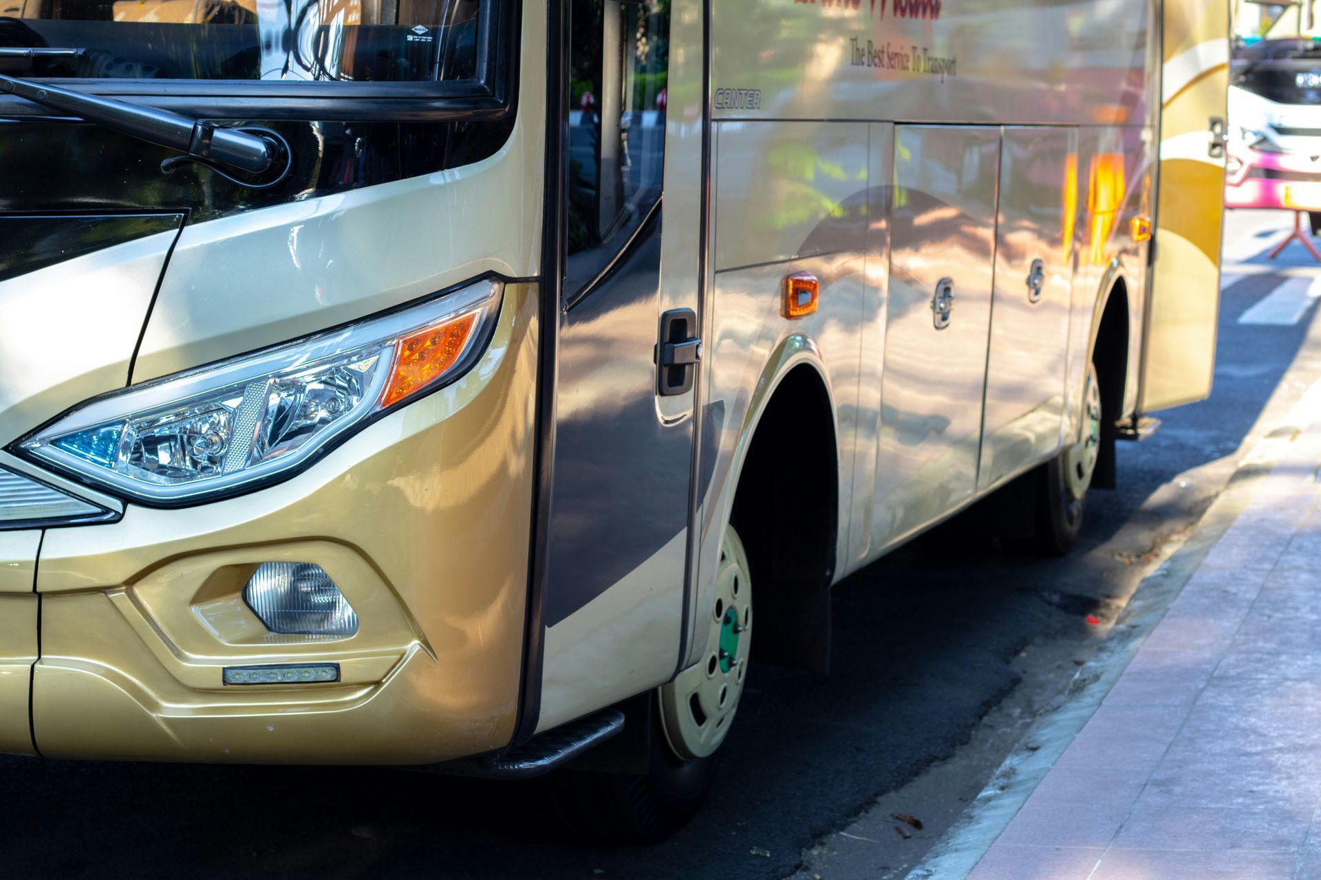 Close-up van een gouden bus geparkeerd langs een stoep. De koplampen en de zijkant van de bus zijn zichtbaar in het zonlicht.