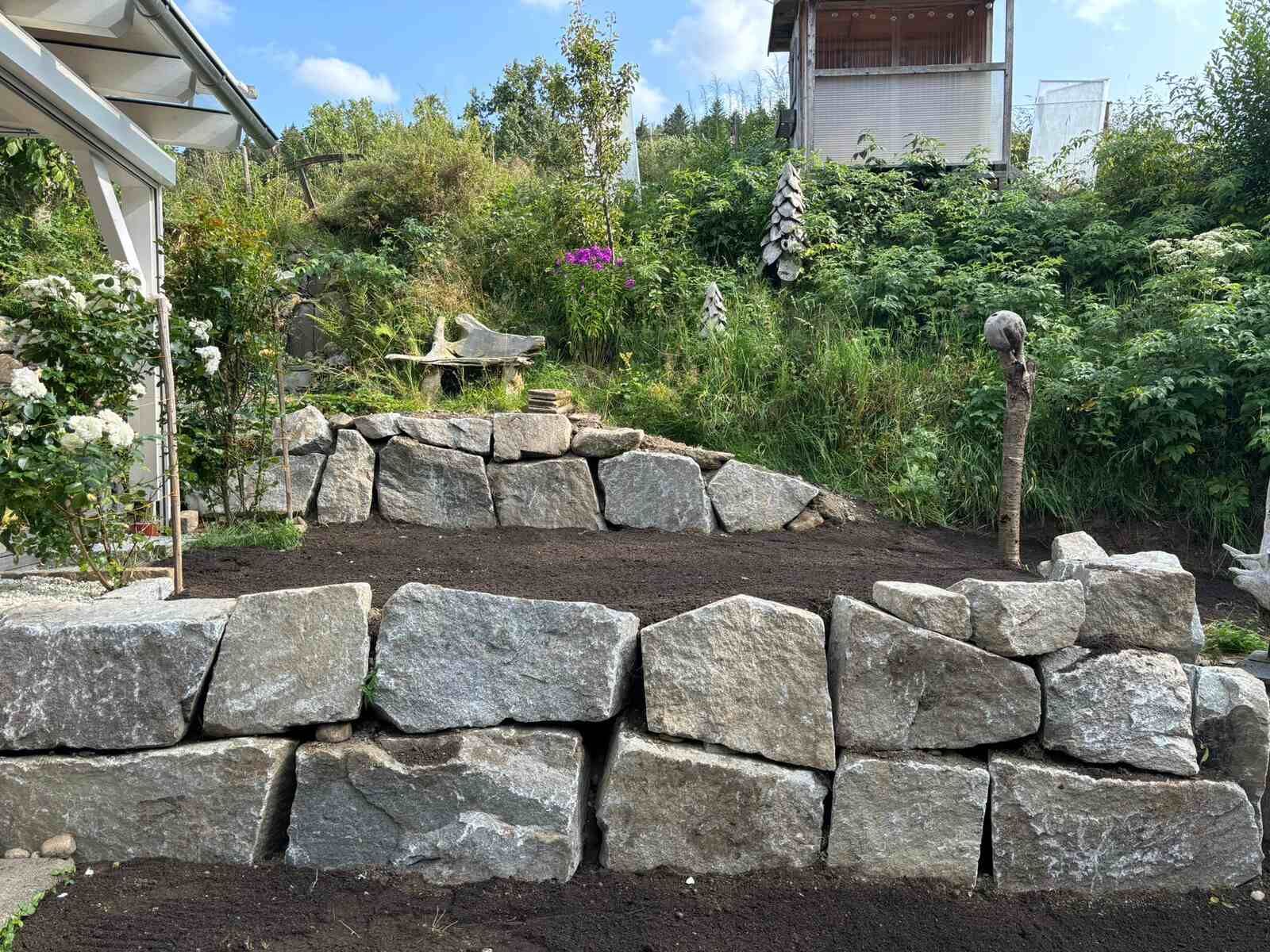 Eine Steinmauer in einem Garten mit einem Haus im Hintergrund.