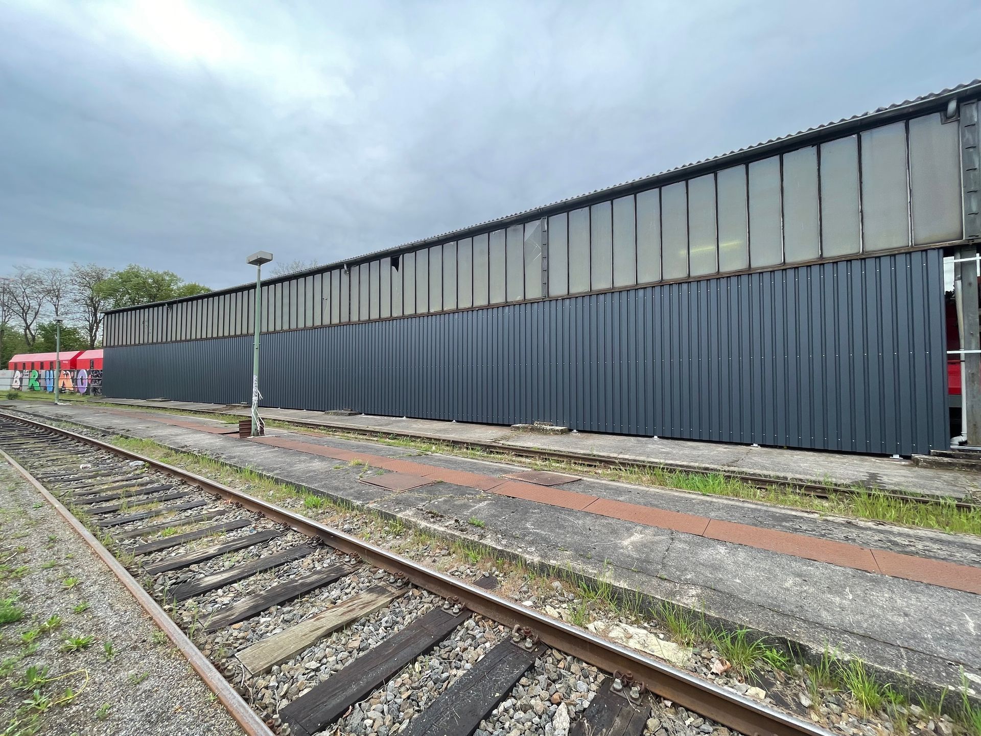 Langer, grauer Wellblechzaun entlang eines stillgelegten Bahngleises unter bewölktem Himmel.