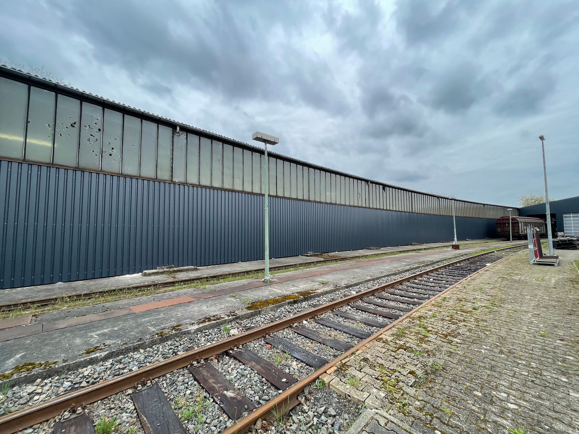 Langer Bahnsteig mit Gleisen, Laternen und einer großen Halle mit Fenstern unter bewölktem Himmel.
