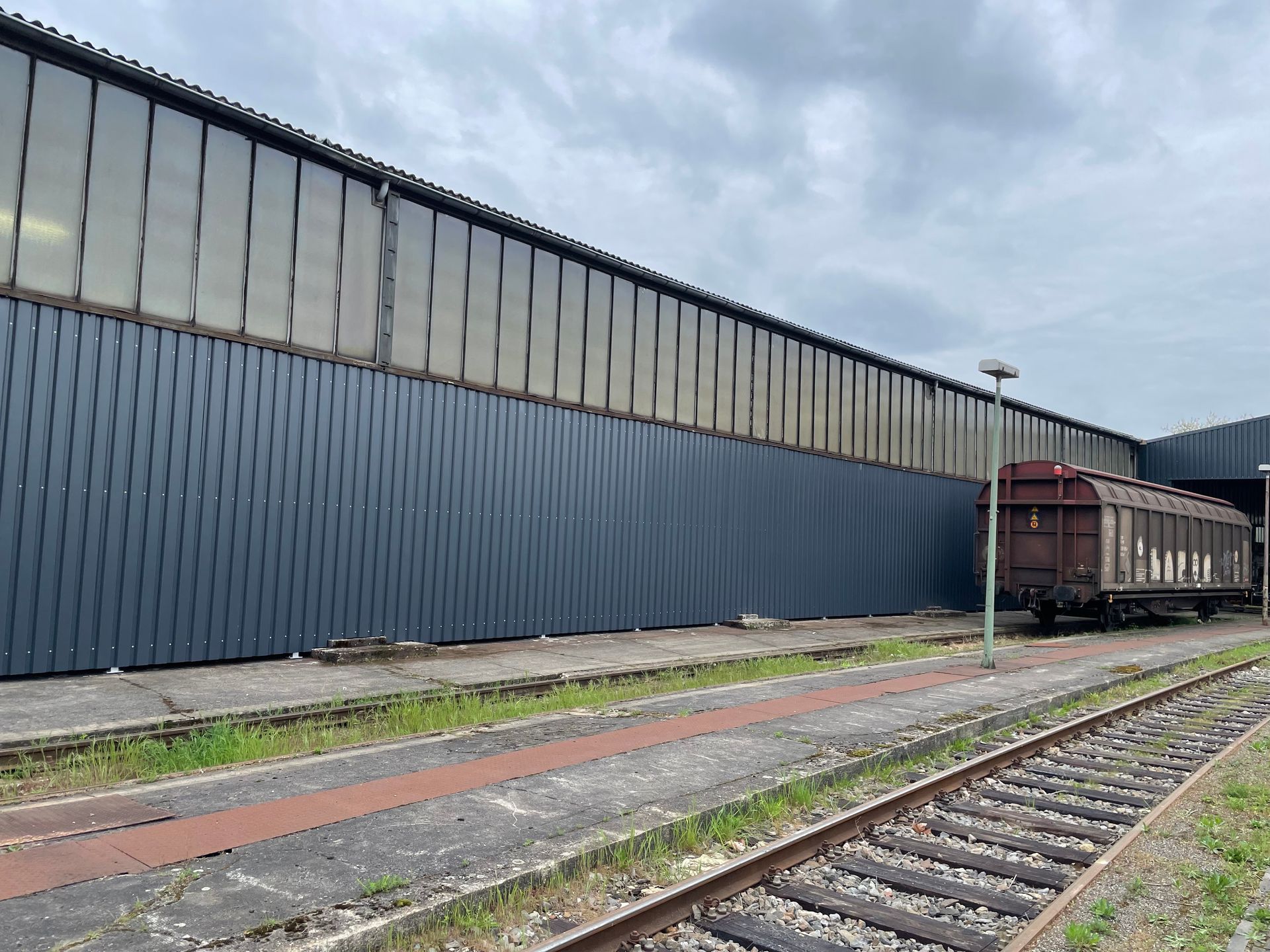 Gleise vor einer langen, blauen Lagerhalle mit einem abgestellten Güterwaggon und einer Straßenlaterne bei bewölktem Himmel.