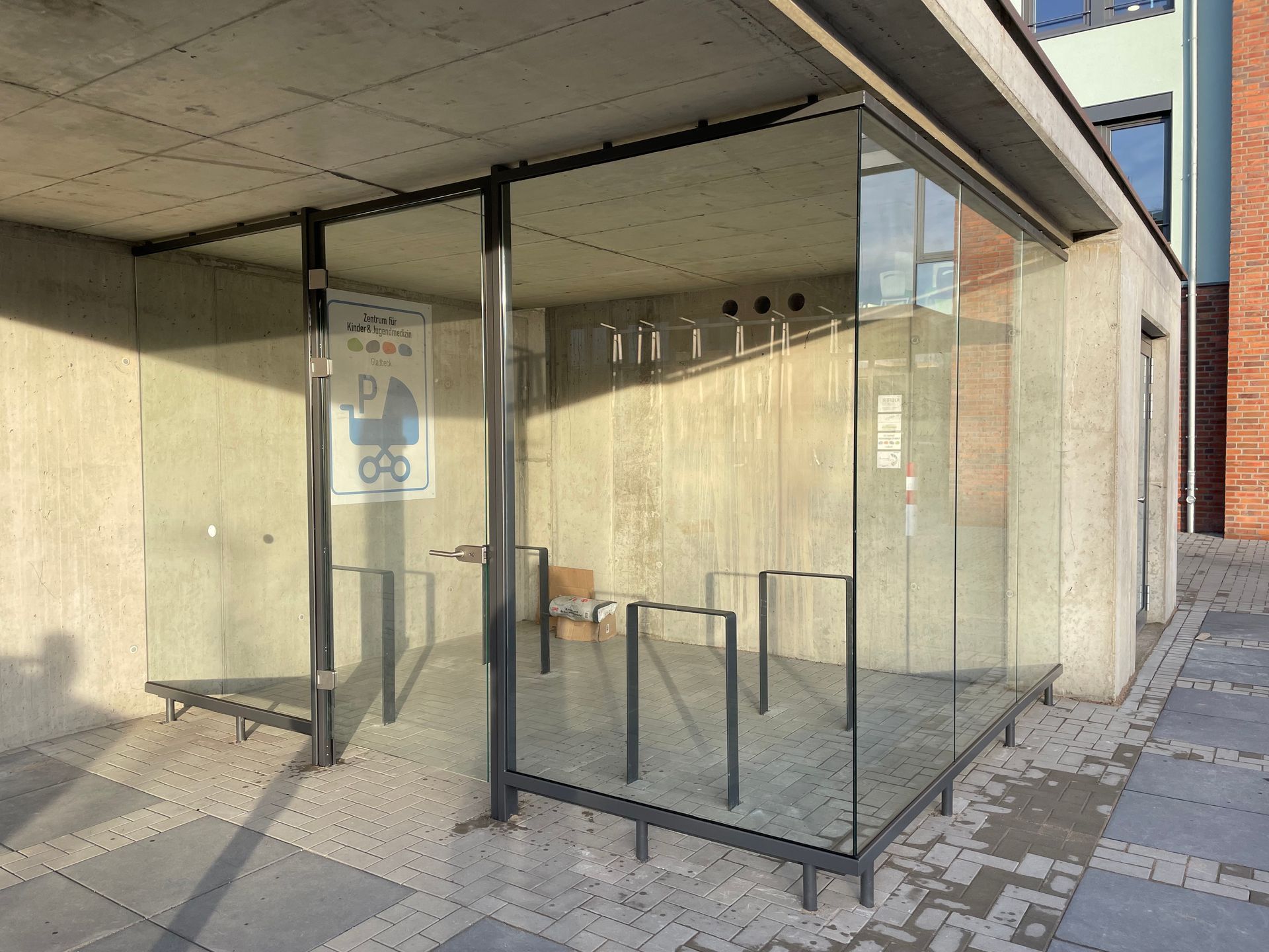 Glasunterstand mit schwarzem Metallrahmen und Fahrradständern auf gepflastertem Boden neben einem Betongebäude.