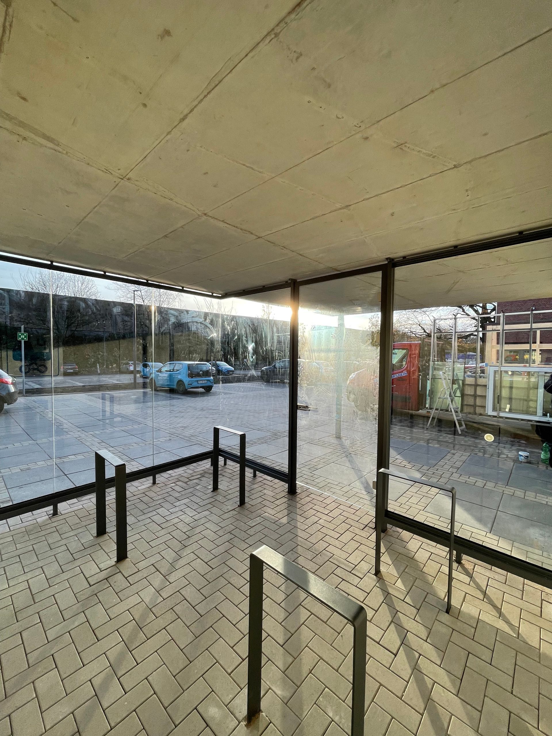 Überdachte Haltestelle mit Glaswänden, Metallgeländern und gepflastertem Boden, im Hintergrund parkende Autos und Gebäude.