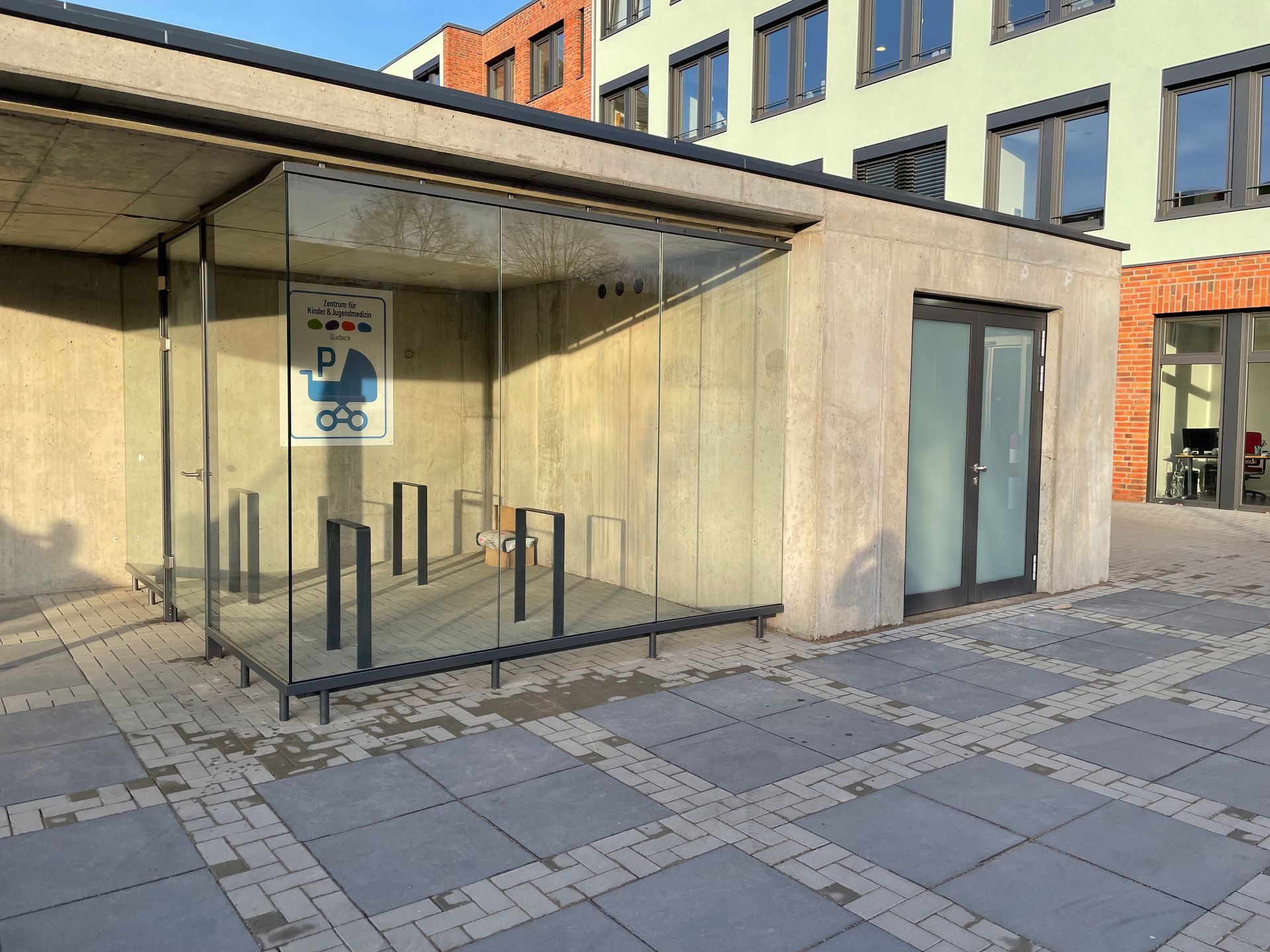 Glasüberdachtes Fahrradparkhaus mit mehreren Fahrradständern vor einem modernen Gebäude aus Beton und Backstein.