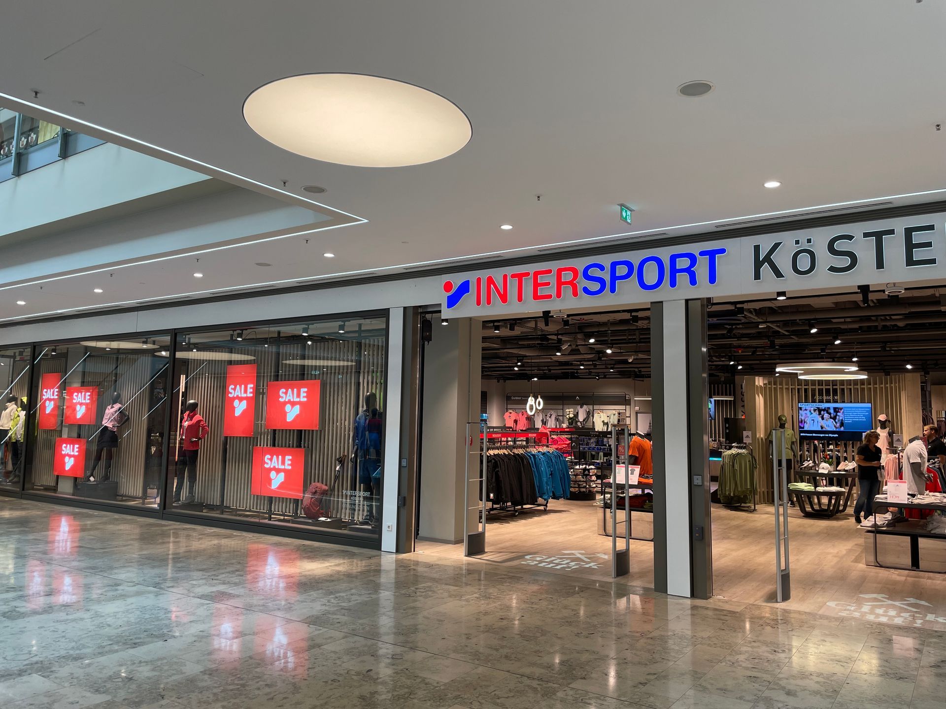 Eingang eines INTERSPORT-Geschäfts mit mehreren roten SALE-Schildern in den Schaufenstern und Sportbekleidung im Inneren.