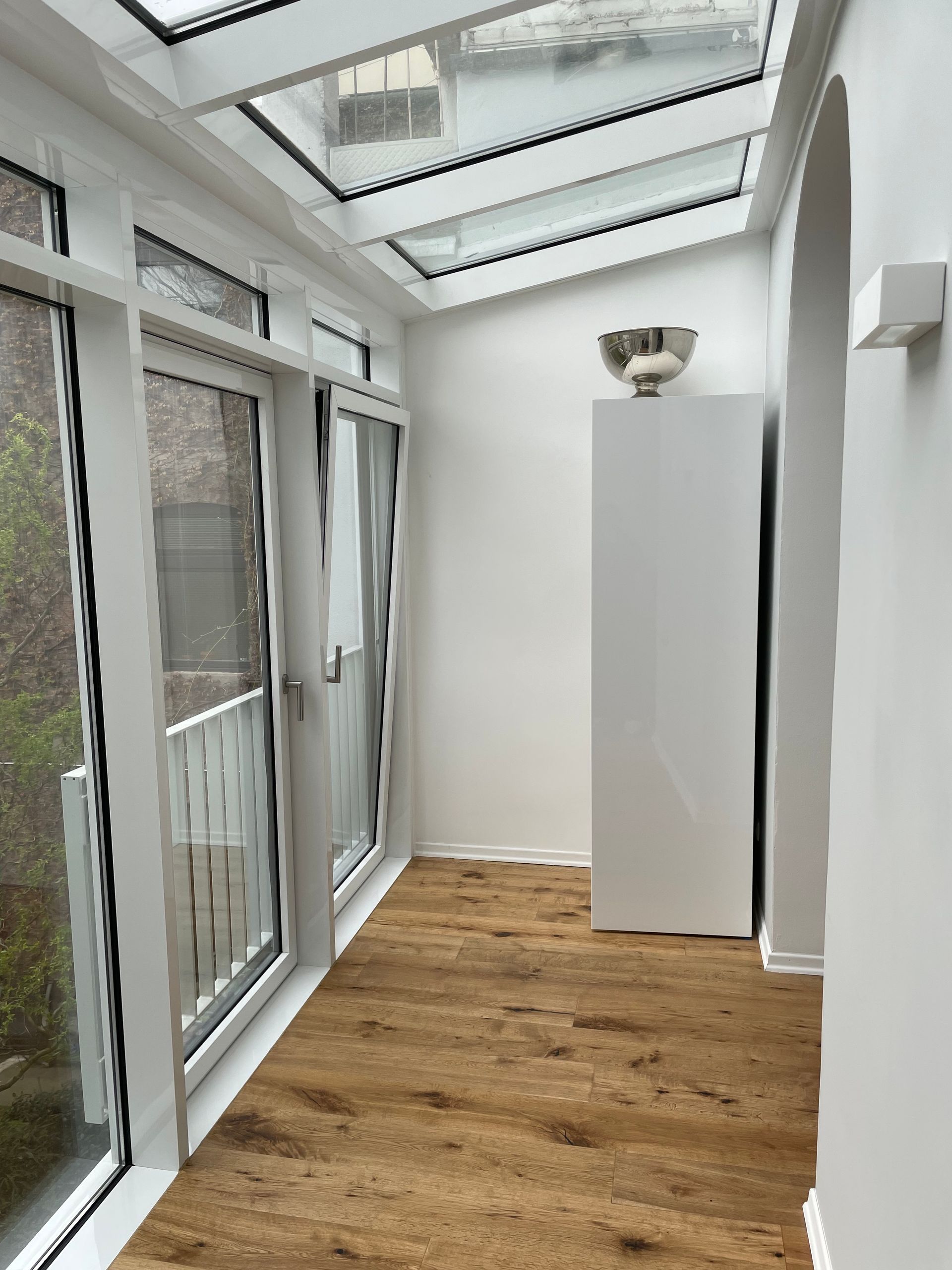 Schmaler heller Raum mit Holzboden, großen Fenstern und einem weißen Schrank mit silberner Schale darauf.