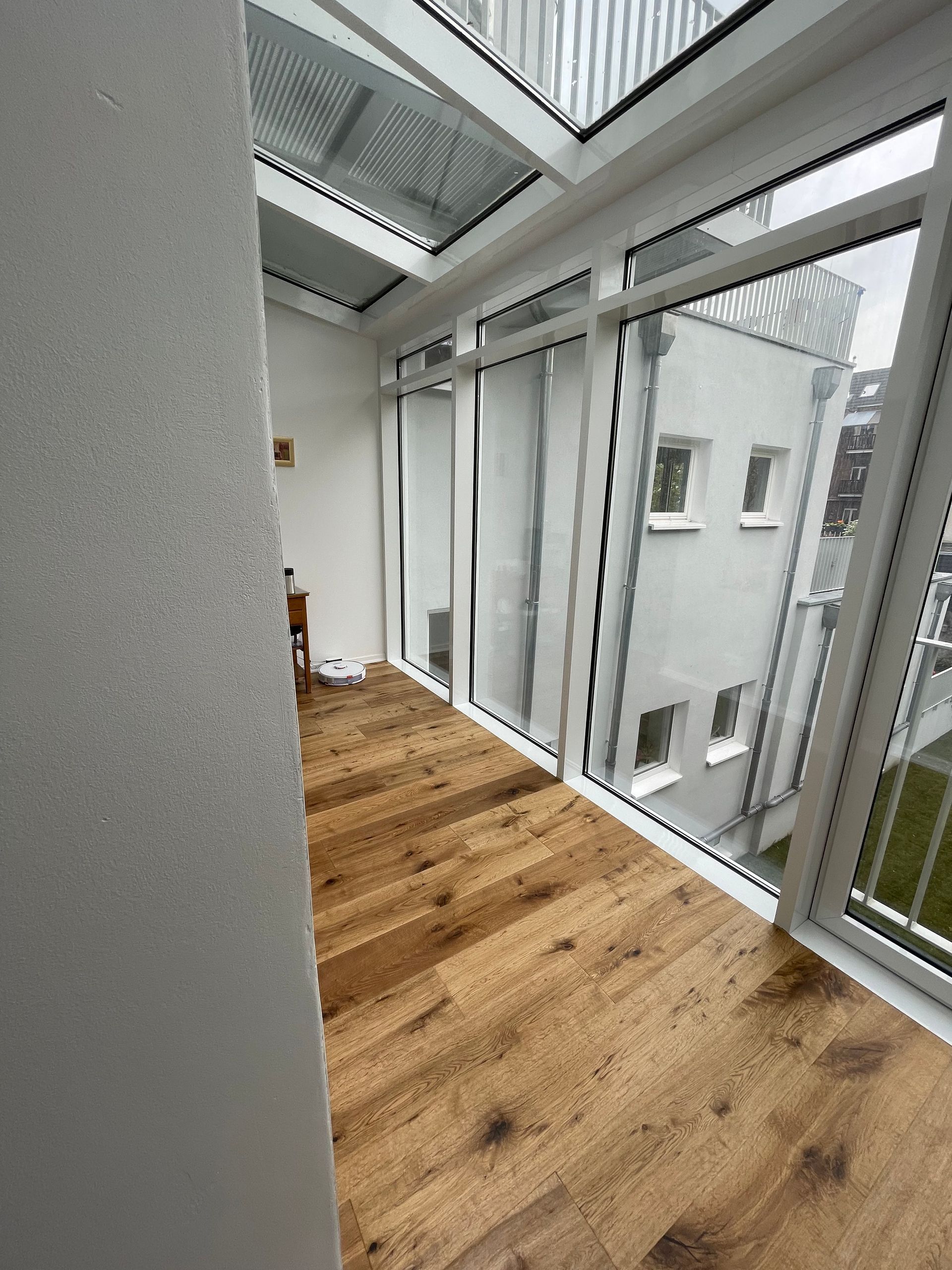 Schmaler Raum mit Holzboden und großen Fenstern, die auf ein benachbartes Gebäude blicken.