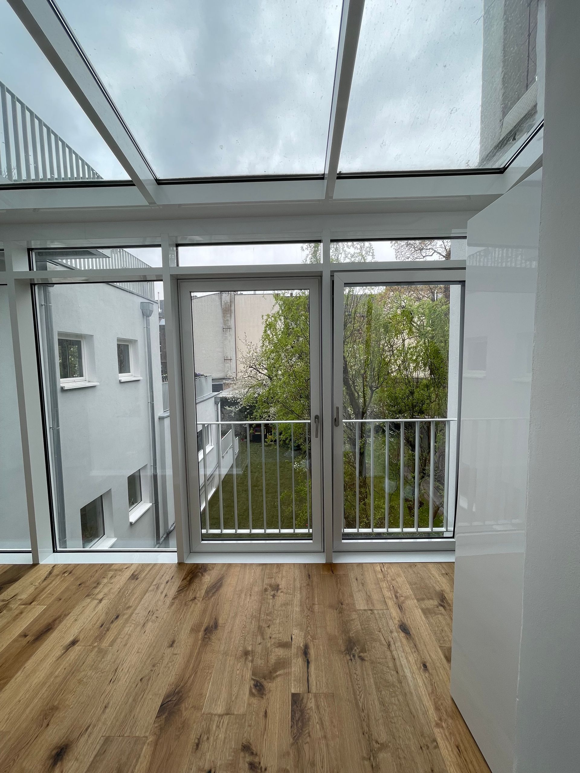 Großes Fenster mit Glasdach und Balkontür, Blick auf grünen Garten und benachbarte weiße Gebäude, Holzboden im Innenraum.
