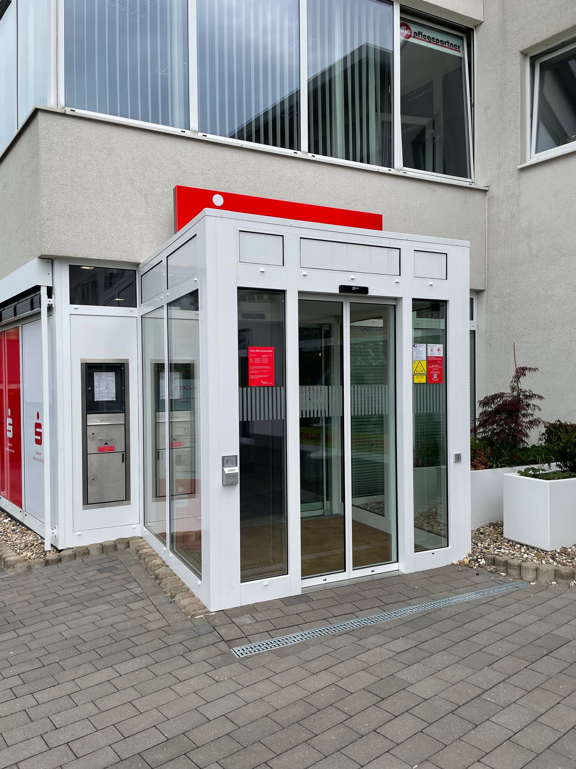 Eingang mit automatischer Schiebetür an einem modernen Gebäude, links ein Briefkasten und rote Schilder sichtbar.