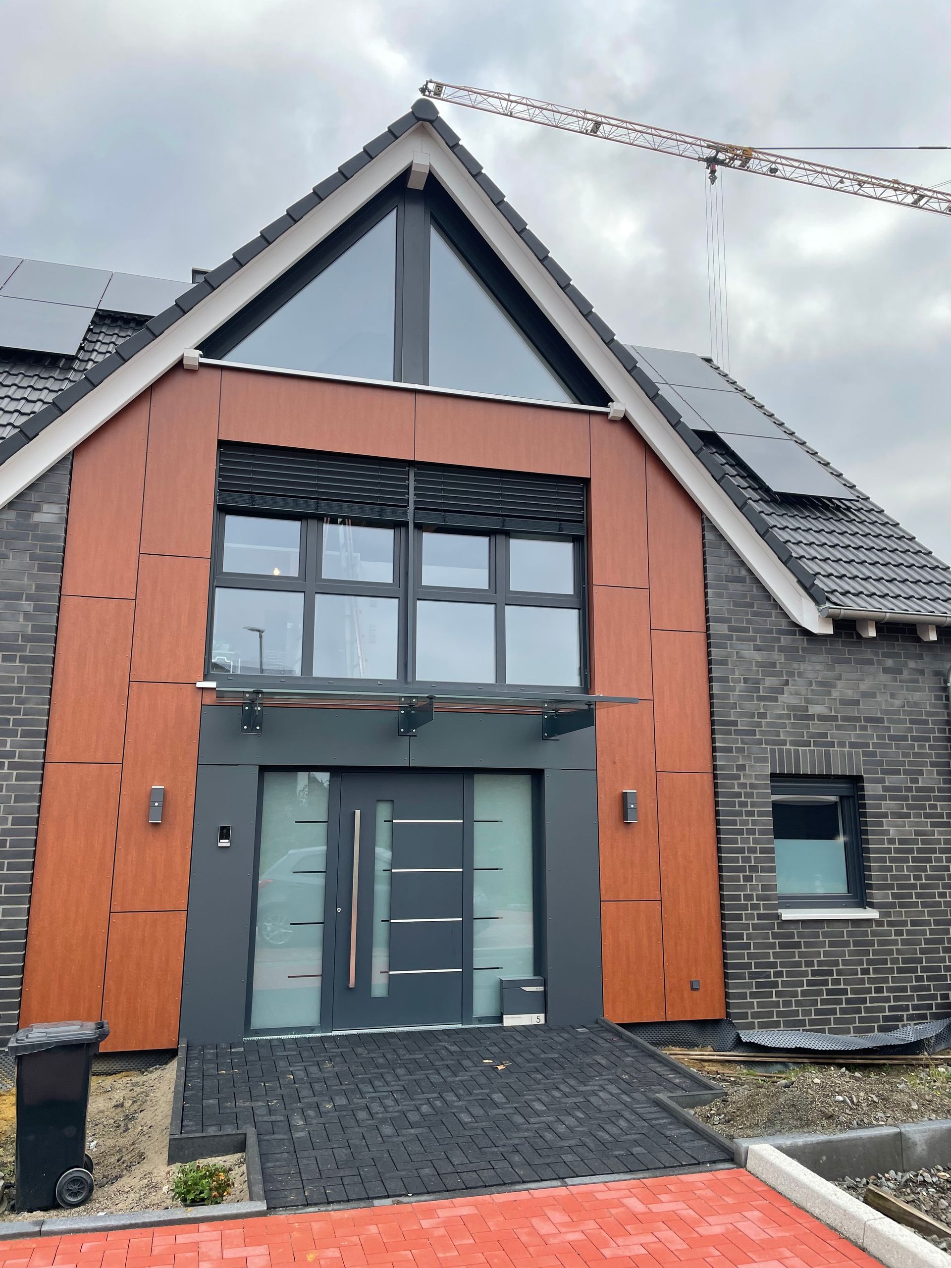 Modernes Haus mit dreieckigem Giebel, Holzverkleidung um die Eingangstür, großen Fenstern und Solarpaneelen auf dem Dach.