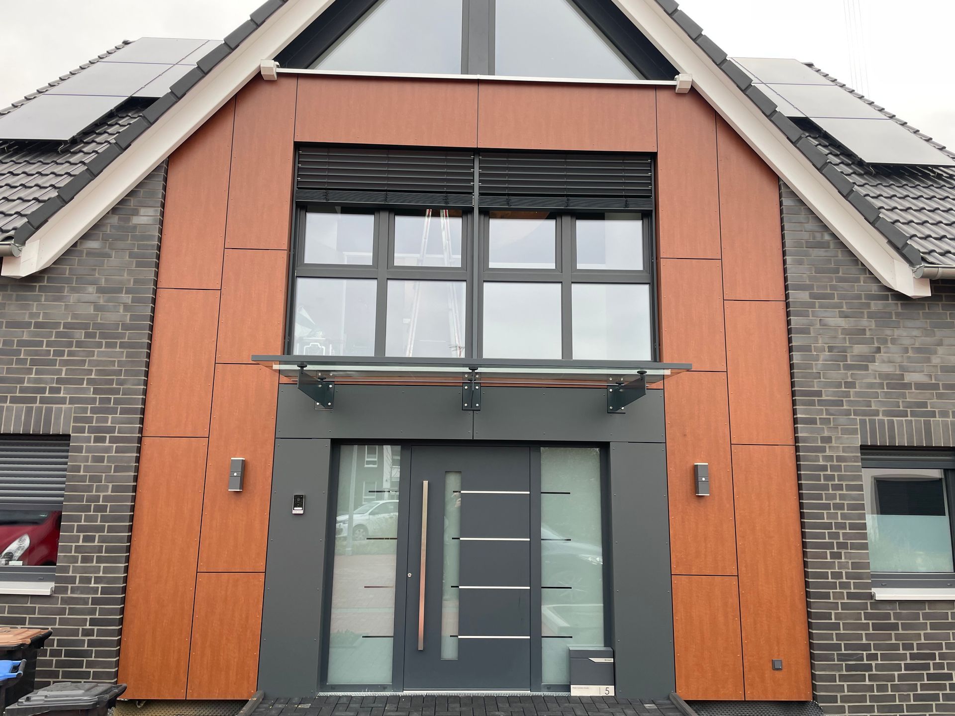 Eingang eines Hauses mit schwarzer Tür, großem Griff, Glasvordach und braunem Fassadenverkleidungselement.