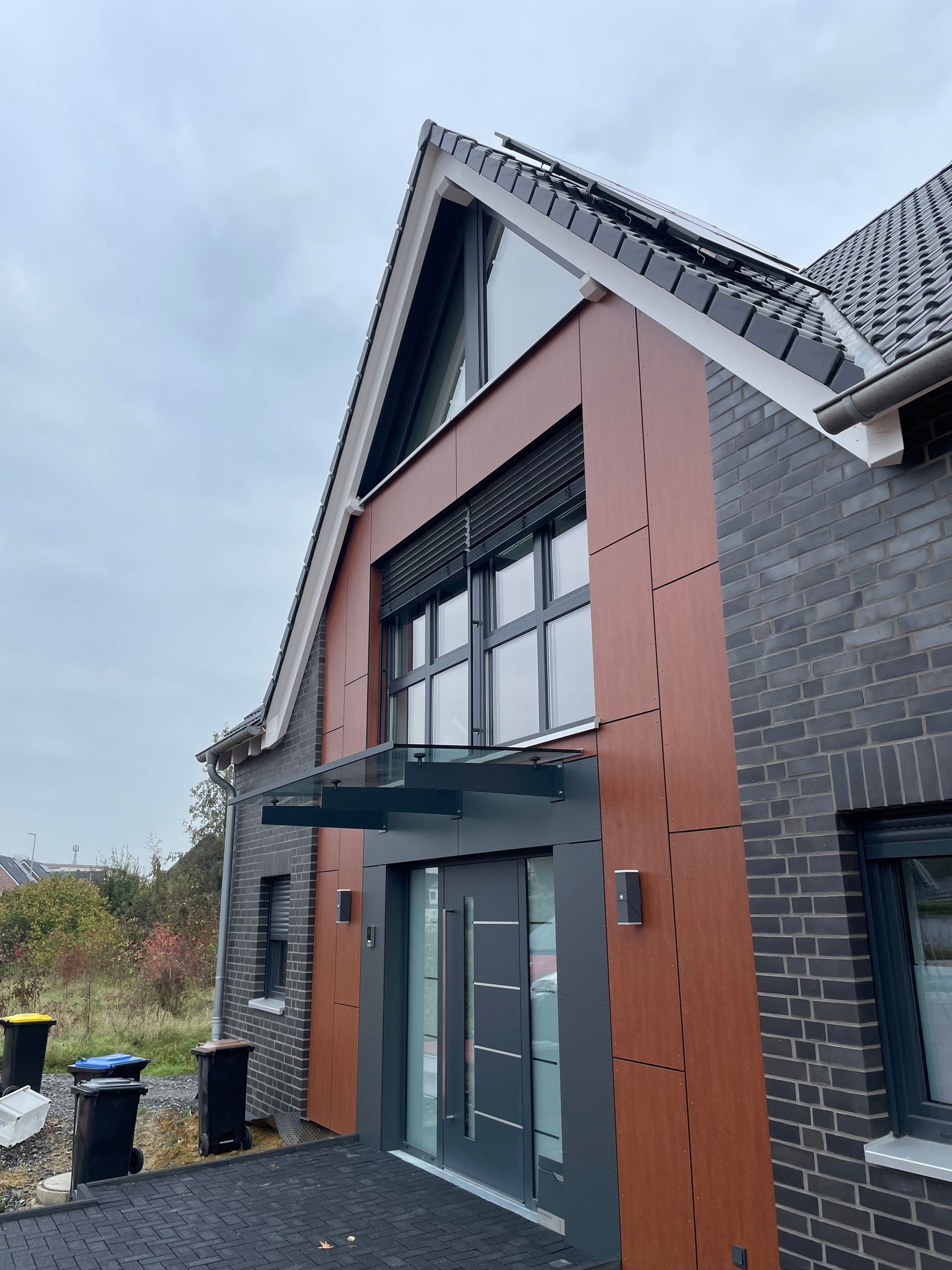 Eingangsbereich eines modernen Hauses mit dunkler Haustür, Glasvordach und braunen Fassadenelementen.