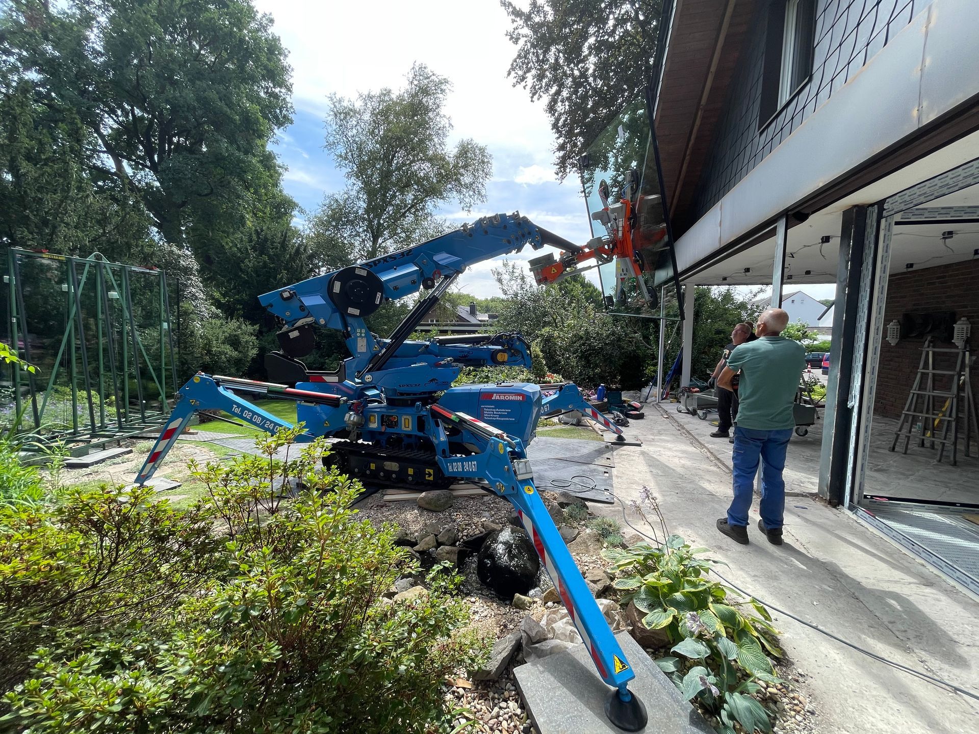 Eine blaue Baumaschine, die im Garten arbeitet, während ein Mitarbeiter eine Glasscheibe