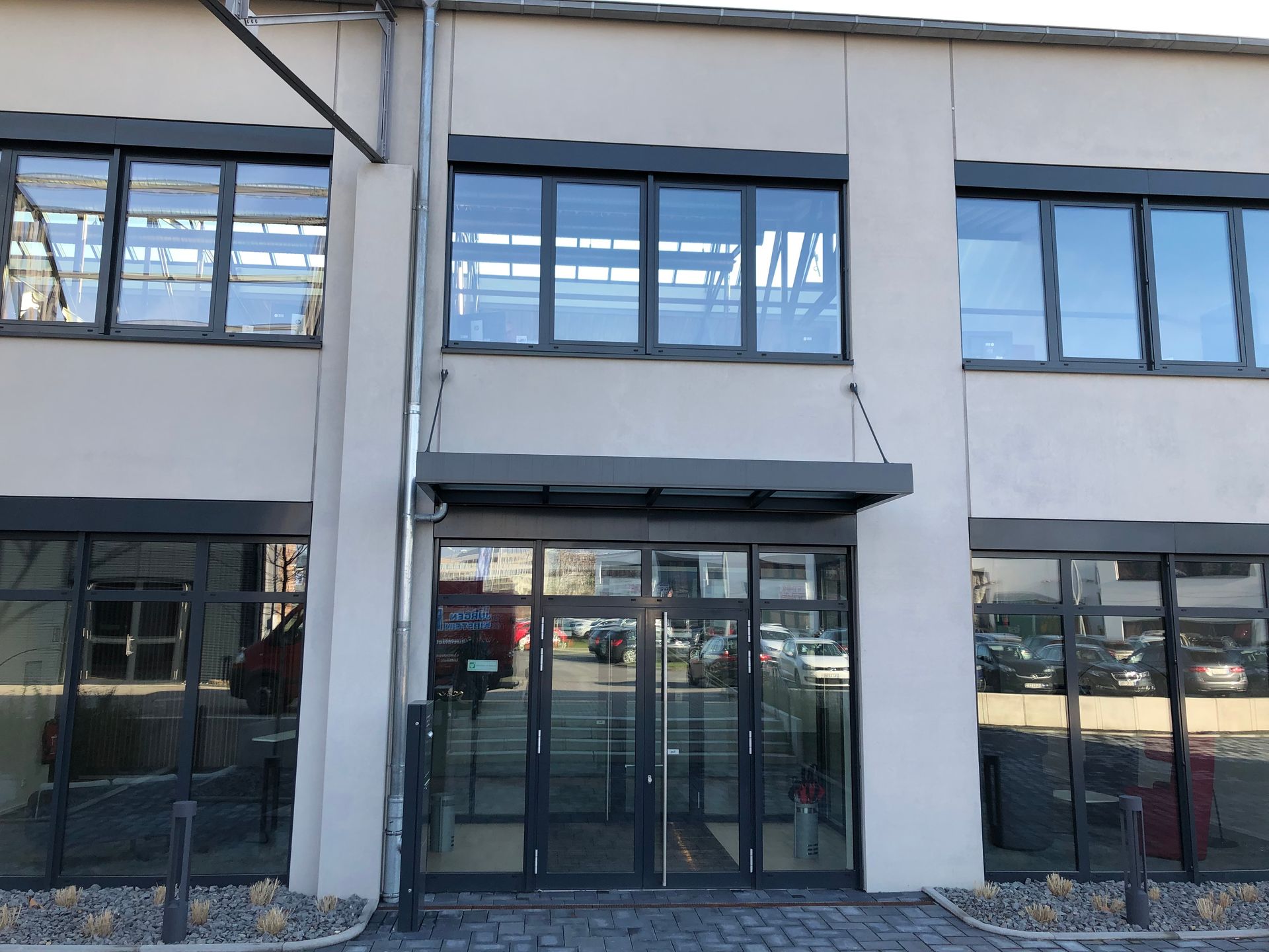 Eingang mit Glastür und zwei großen Fenstern darüber an einem hellen Gebäude, Spiegelungen von Autos auf der Glasfläche.