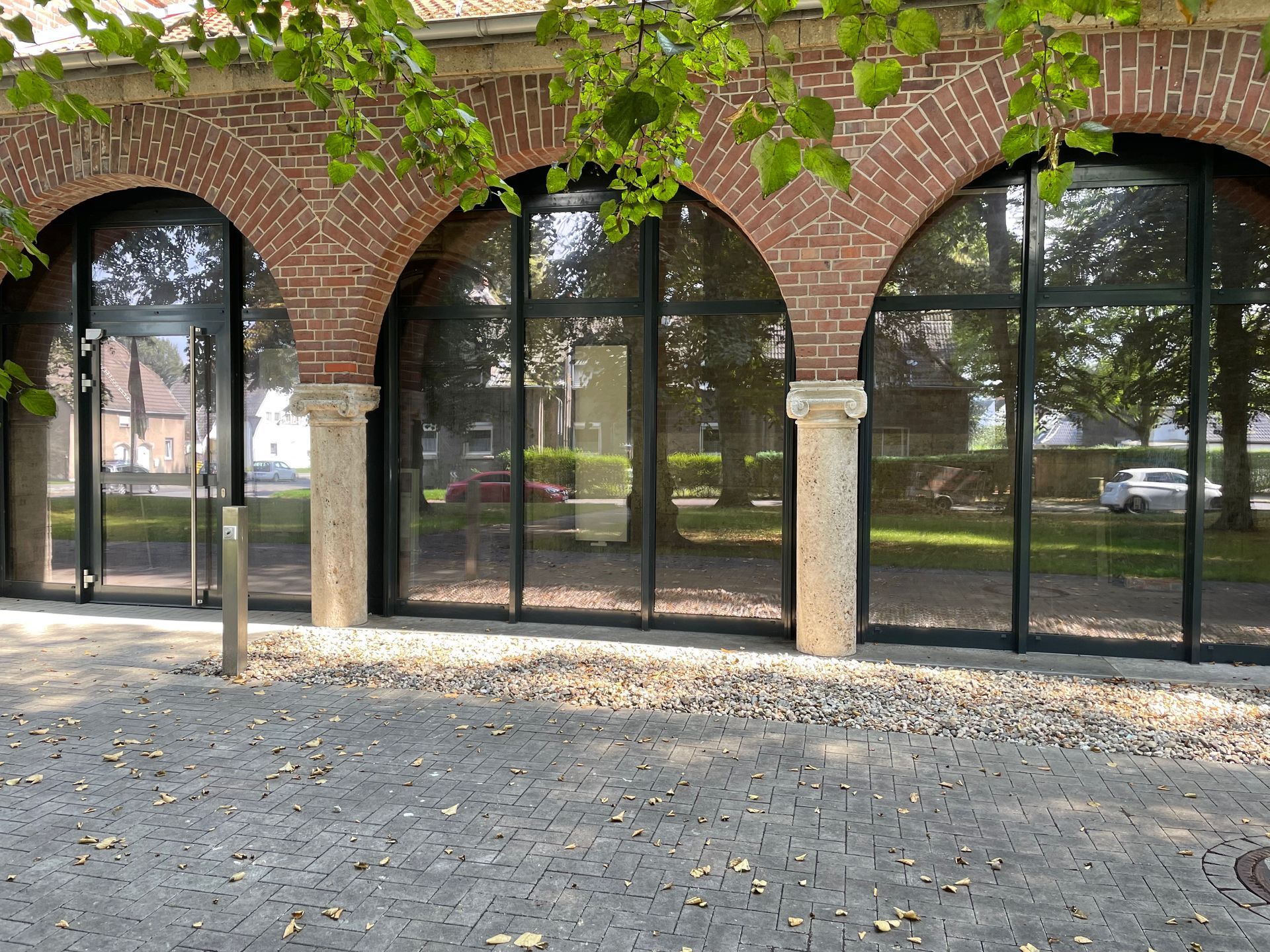 Backsteinfassade mit drei großen, schwarzen Glasbögen und zwei steinernen Säulen, davor gepflasterter Boden mit Laub.