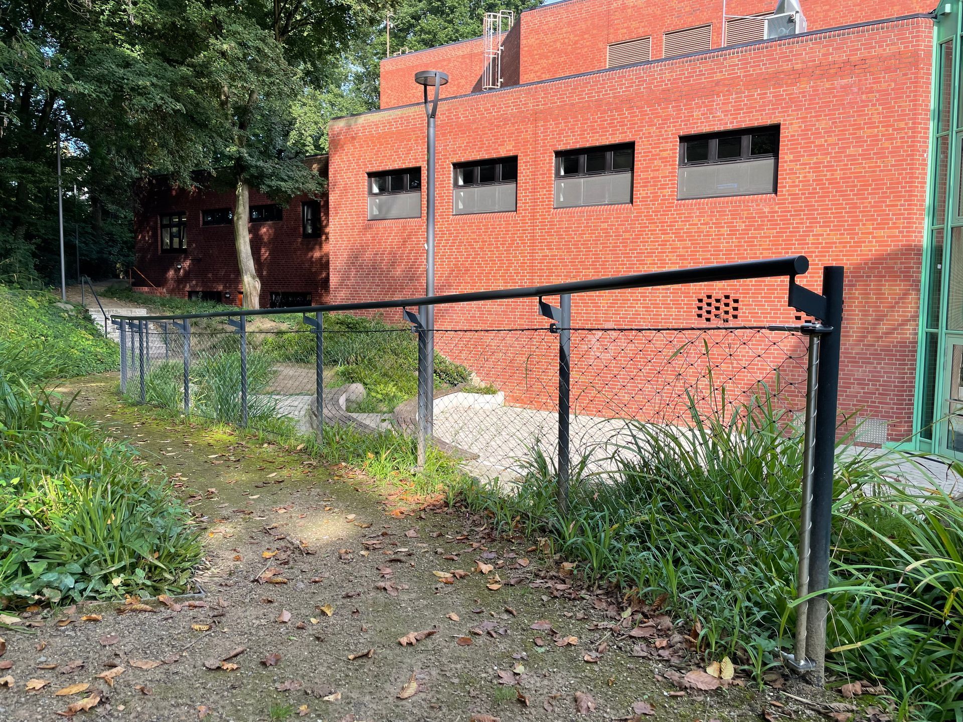 Schräg verlaufender Weg mit Metallgeländer, umgeben von Gras und Büschen, im Hintergrund rotes Backsteingebäude mit Fenstern.