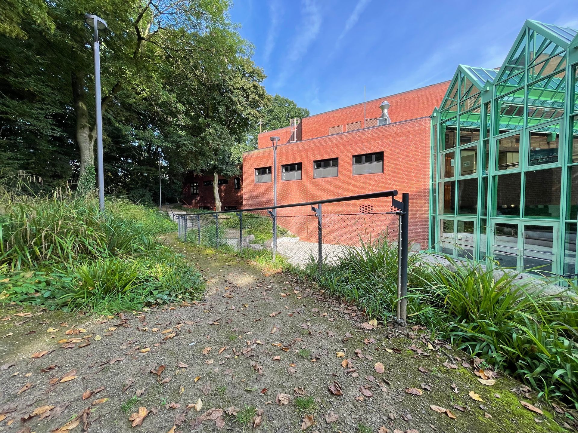 Schmaler Weg mit Laub, rechts ein grünes Glasdach und ein rotes Backsteingebäude, links Bäume und Gras.