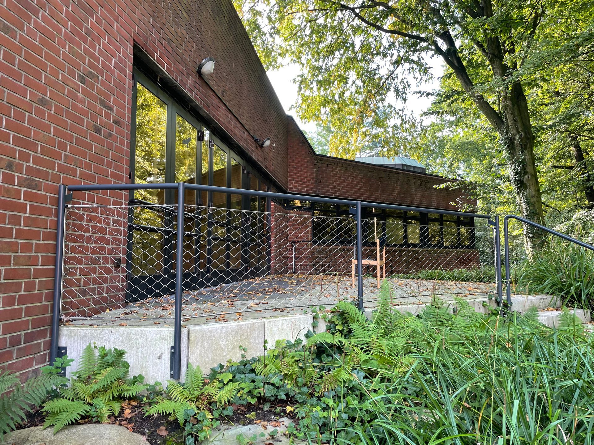 Backsteinwand mit großen Fenstern, davor eine Terrasse mit schwarzem Metallgeländer und Pflanzen im Vordergrund.