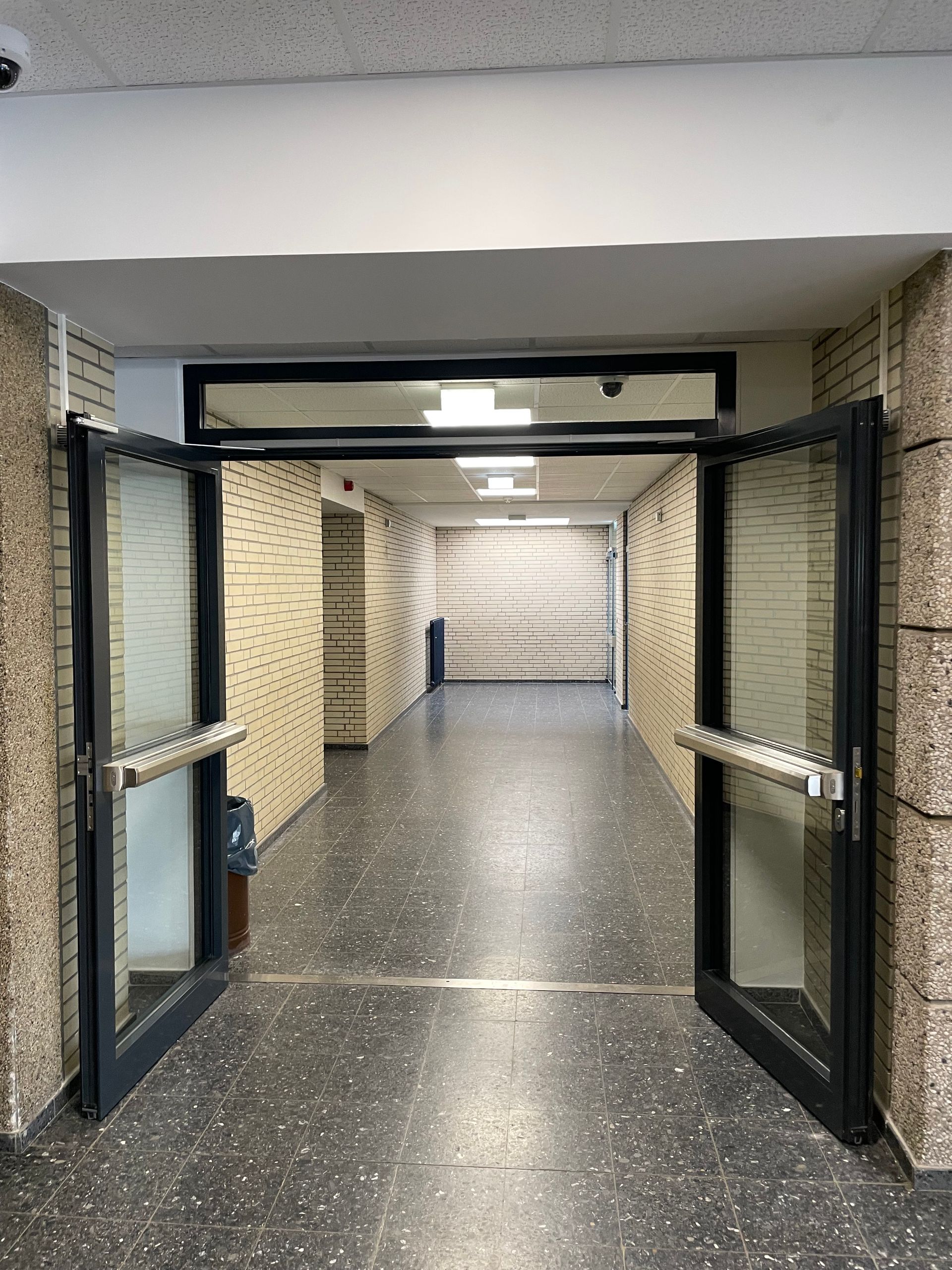 Offene doppelflügelige Glastür mit schwarzen Rahmen führt in einen langen Flur mit beigen Klinkerwänden und dunklem Boden.