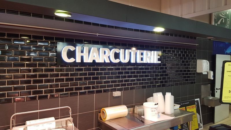 Enseigne Charcuterie sur un mur carrelé de noir, éclairée par des plafonniers. Comptoir en acier inoxydable en dessous.