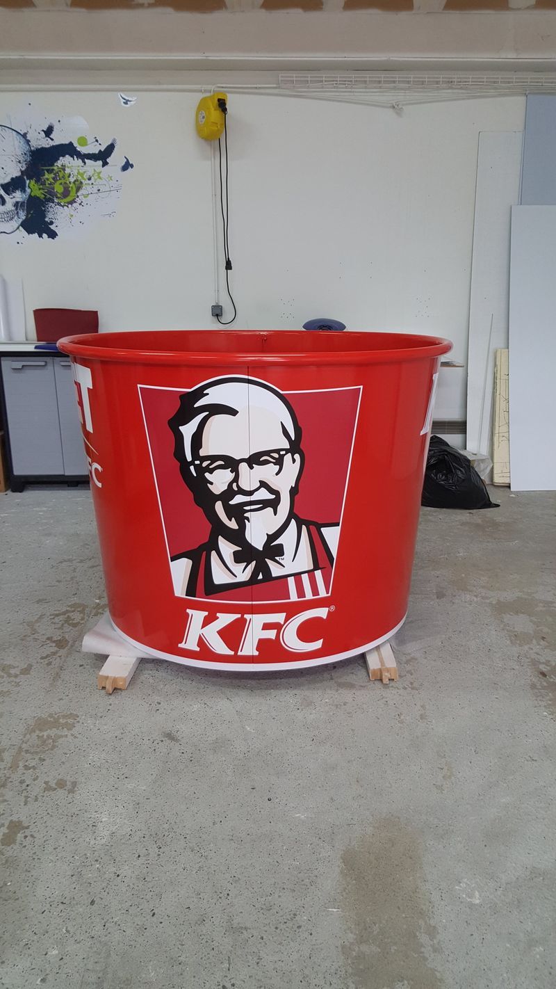 Grand seau rouge KFC orné du logo du Colonel Sanders, posé sur des supports en bois dans un studio.