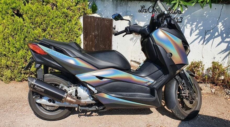 Un scooter Yamaha noir mat aux reflets irisés et changeants est garé sur un chemin de gravier, adossé à un mur blanc.